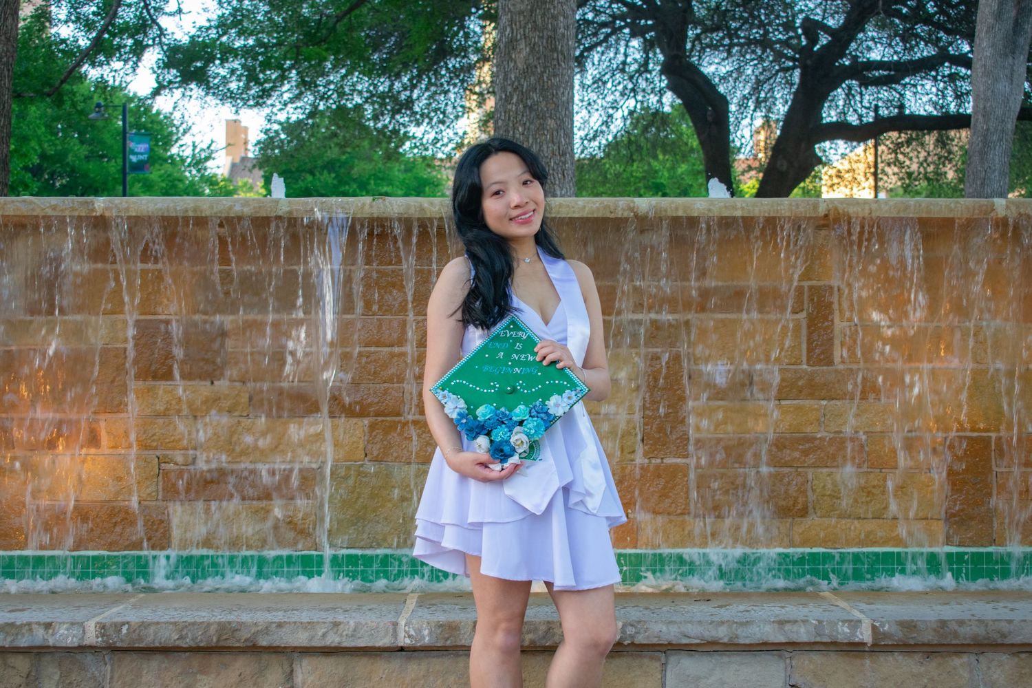 Young woman in white dress holding decorated graduation cap poses outdoors near stone fountain wall.