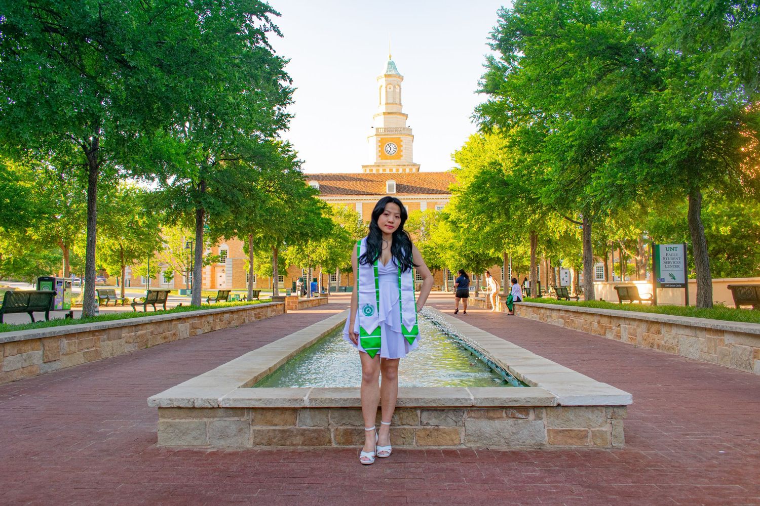 Graduate in white regalia poses by a fountain on a brick plaza with lush trees and a historic building at Baylor University.