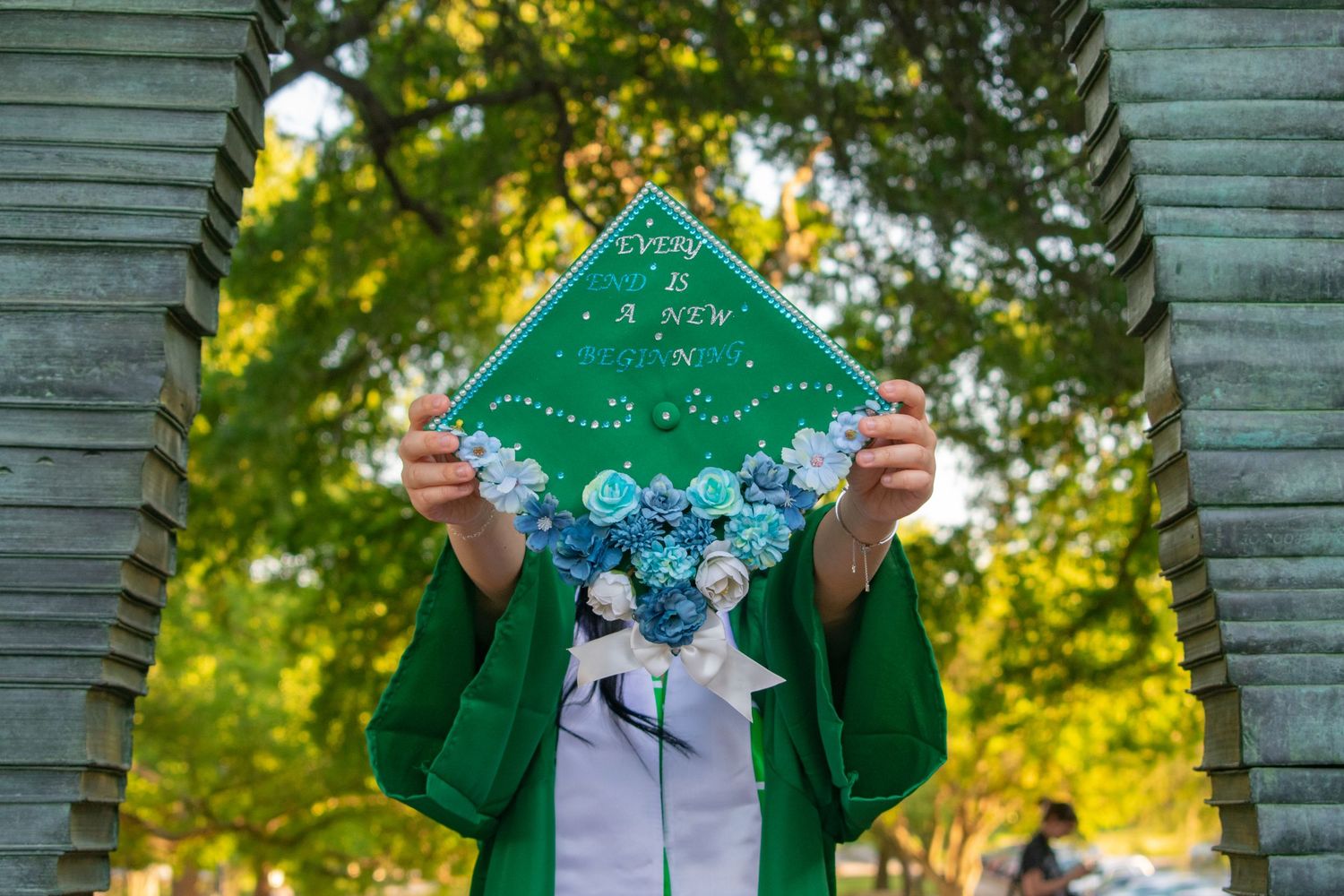 Graduate holds decorated green cap with floral design under stone arch at outdoor graduation ceremony.