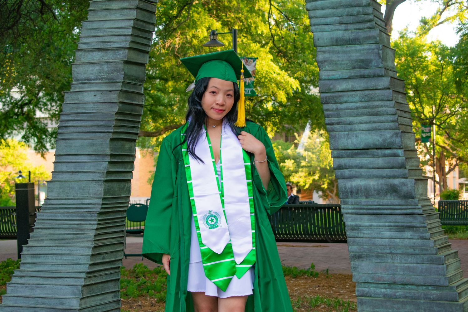 Young woman in green graduation cap and gown poses between stone pillars on a university campus.
