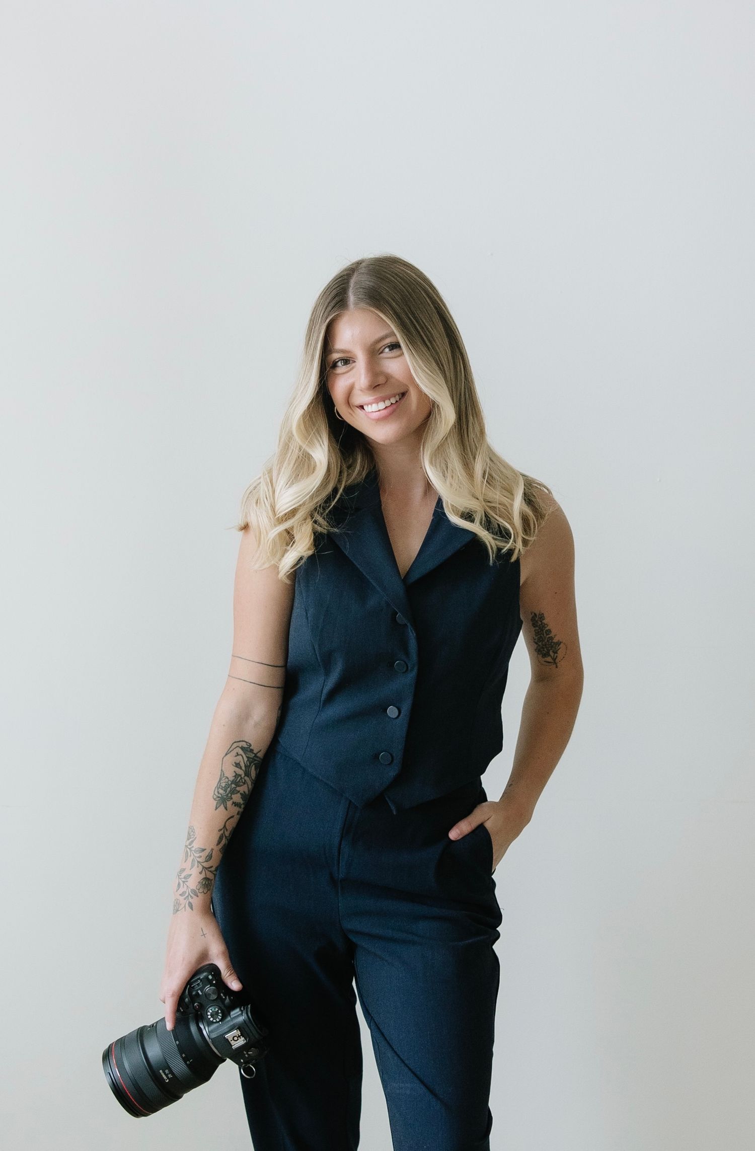 Smiling blonde photographer with tattoos holds camera wearing navy jumpsuit.