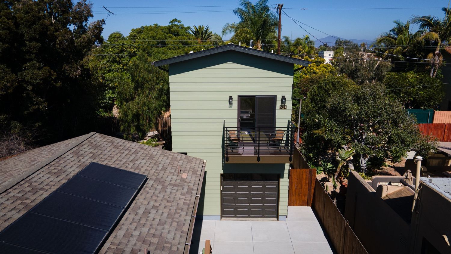 Close-up exterior of a modern green two-story ADU with garage, balcony, and mountain views behind.