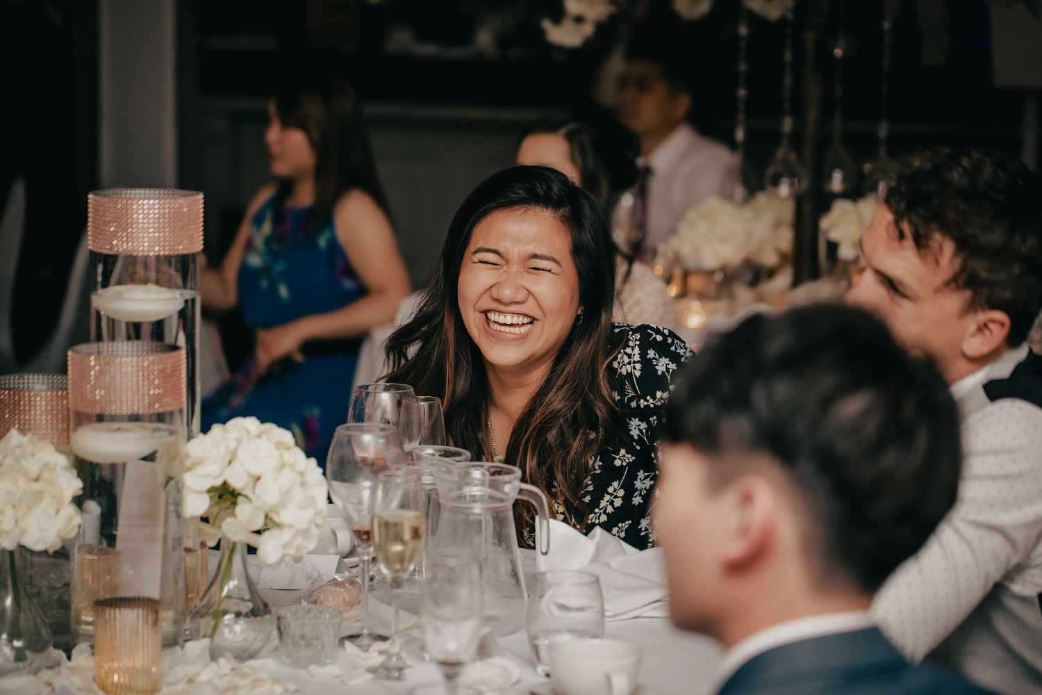 Wedding guest laughing joyfully at decorated reception table with white floral centerpieces.