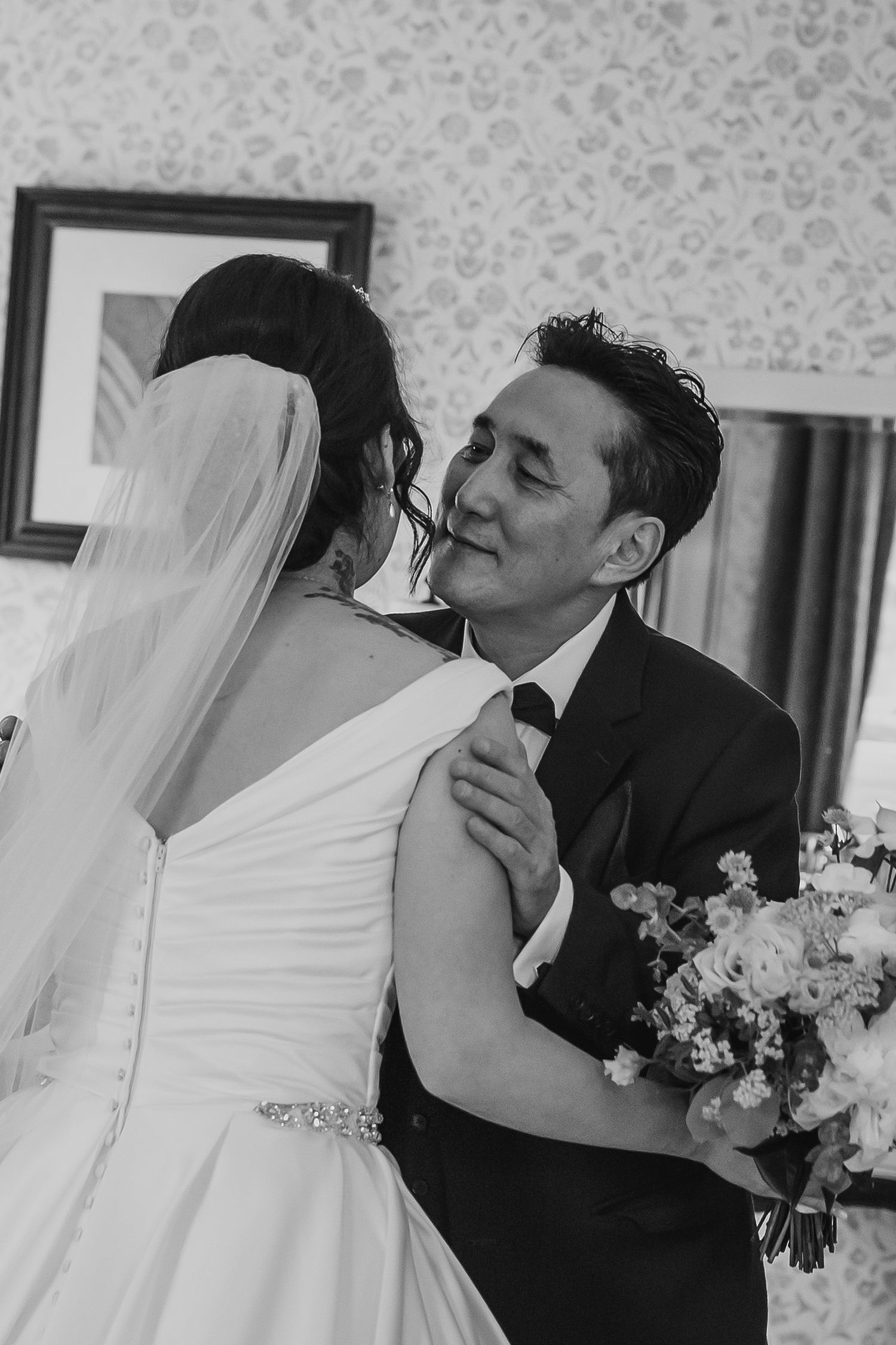 A groom smiles warmly while embracing his bride in a white gown in black and white photo.