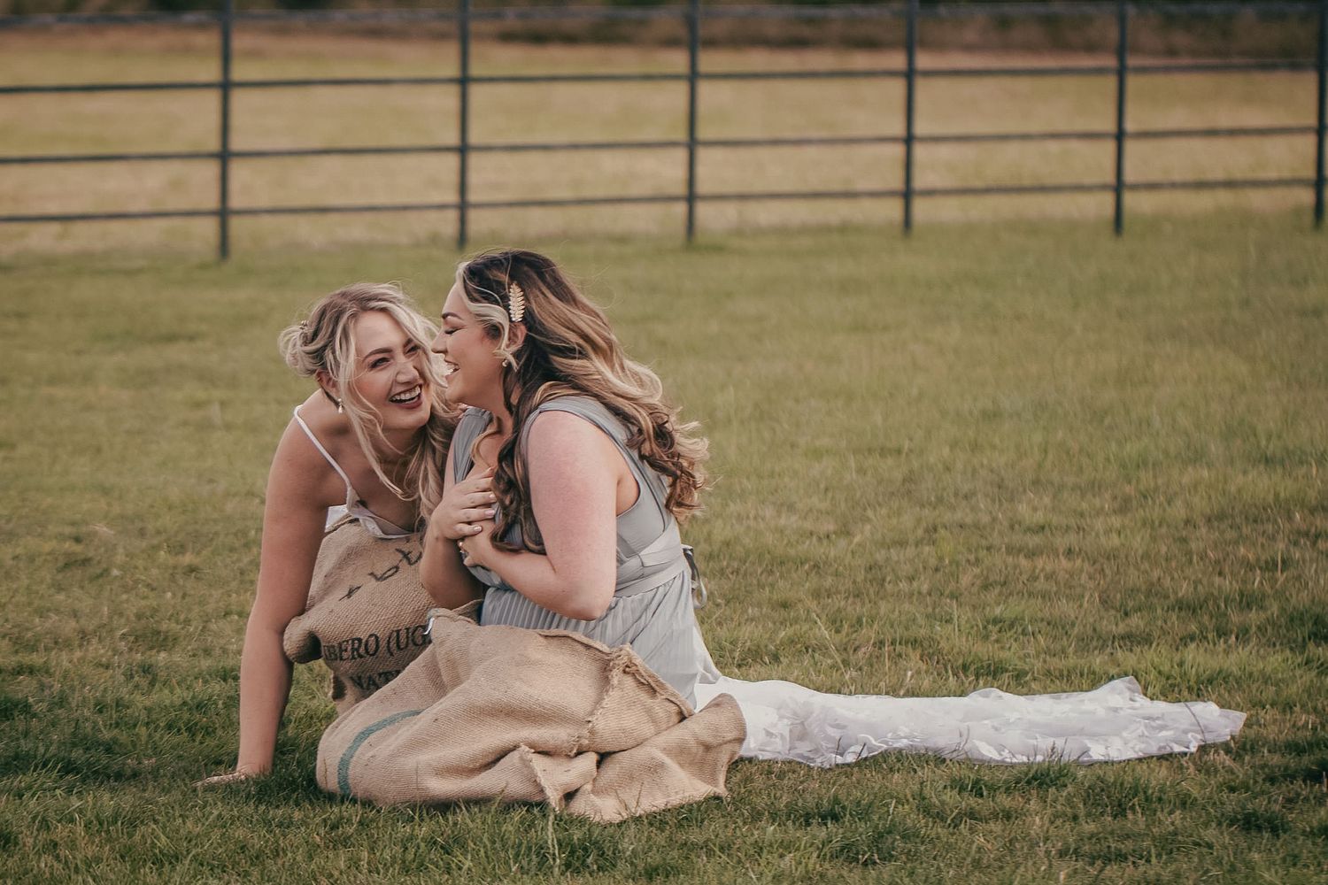 Bride and bridesmaid sharing joyful moment sitting together in burlap sacks on wedding day.
