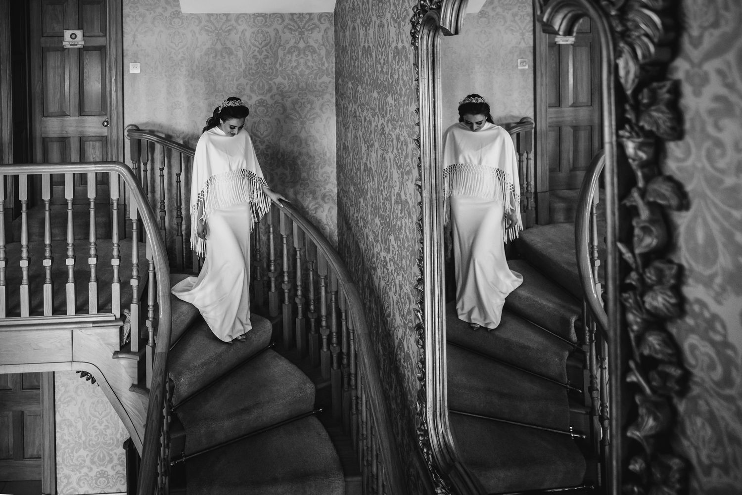 Elegant bride in long white gown descends grand staircase reflected in ornate vintage mirror.