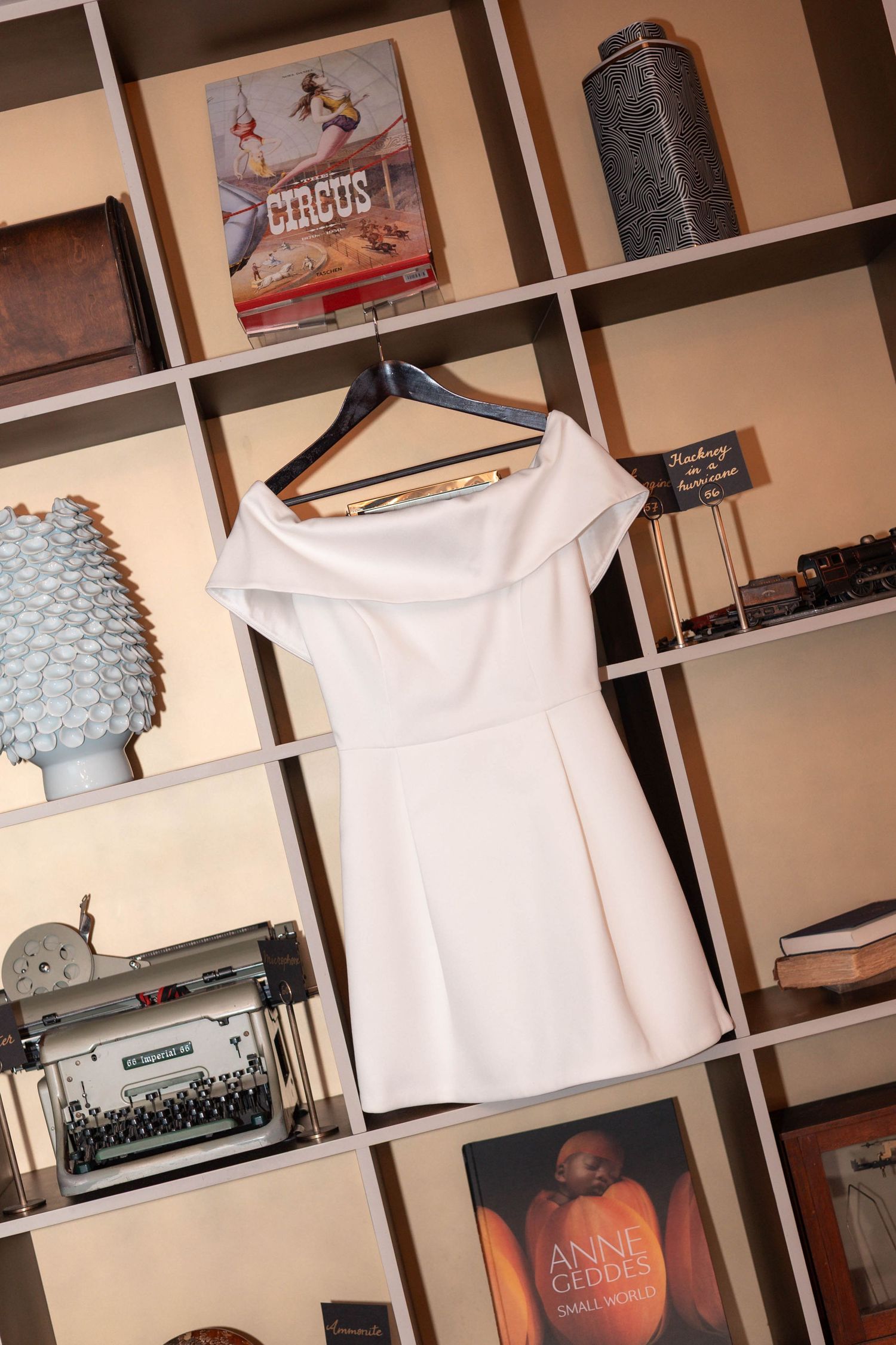 White off-shoulder mini dress on hanger displayed against decorative shelving with books and vintage typewriter.
