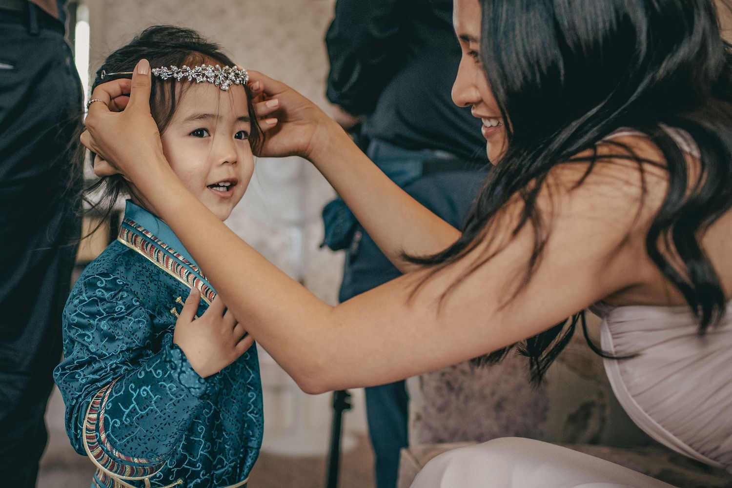 A woman places a floral crown on a young girl in a blue dress at a wedding celebration.