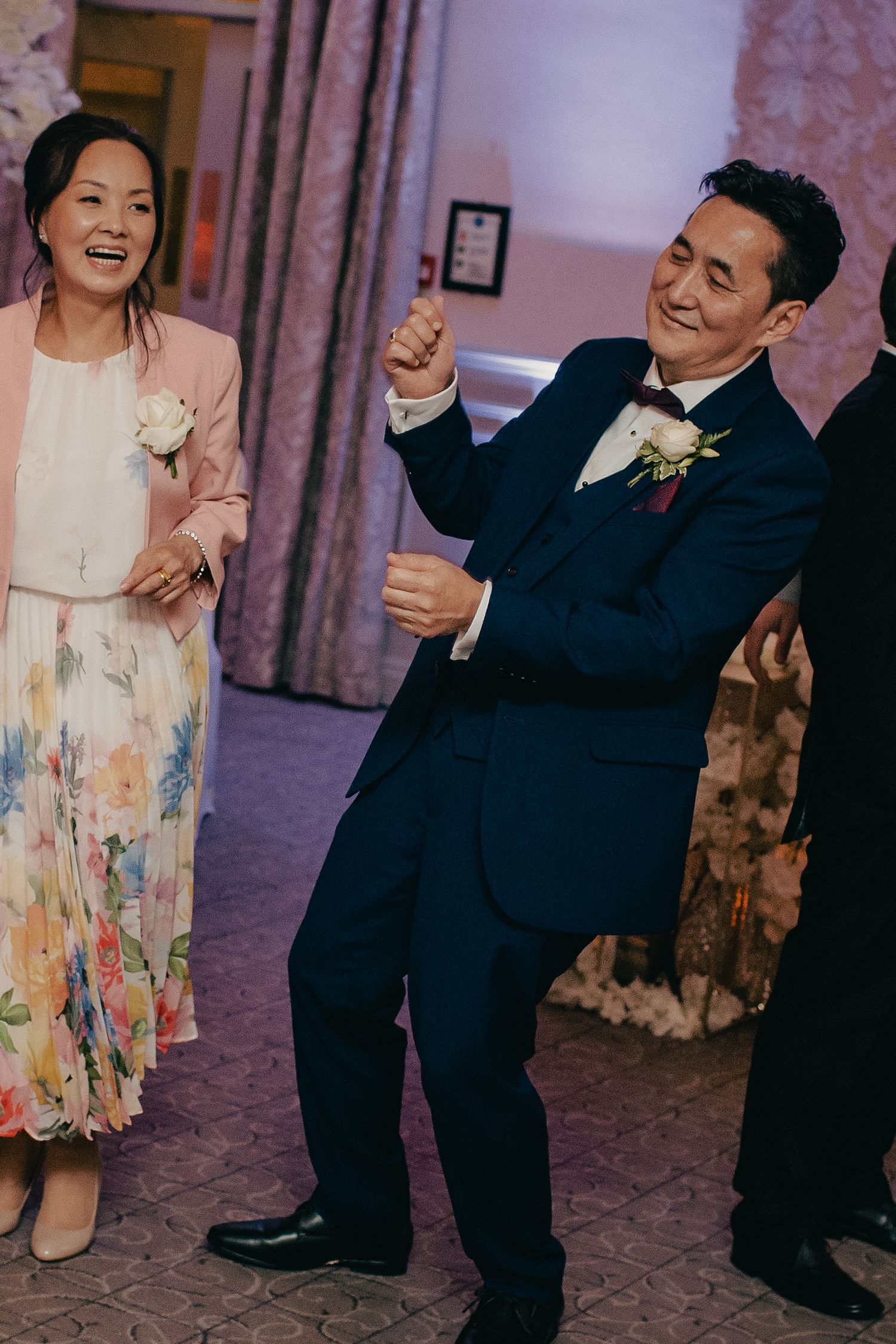 Groom in navy suit dancing enthusiastically with smiling woman in floral dress at wedding reception.