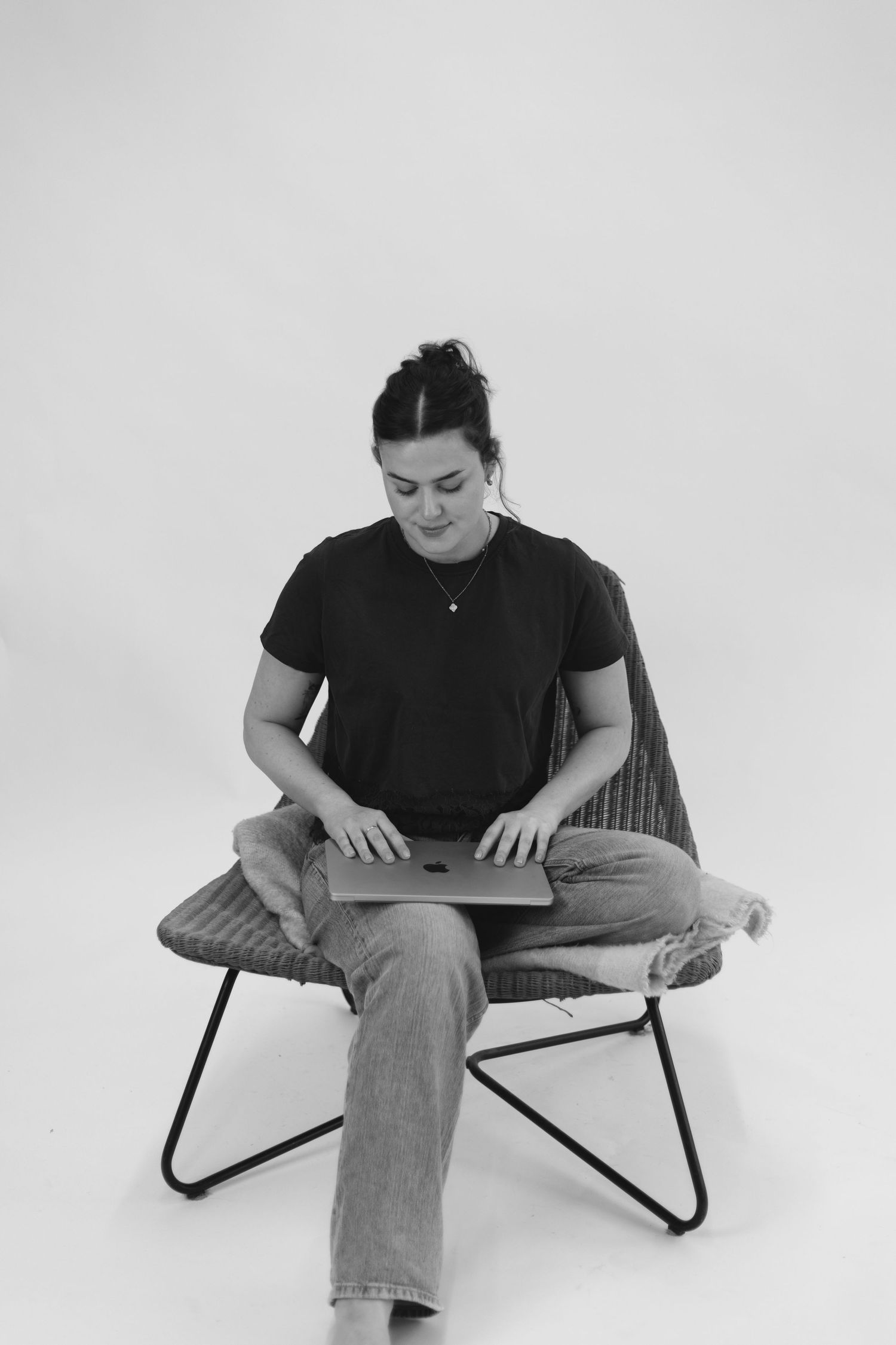 Young woman with hair up sits on modern chair typing on Apple MacBook laptop.