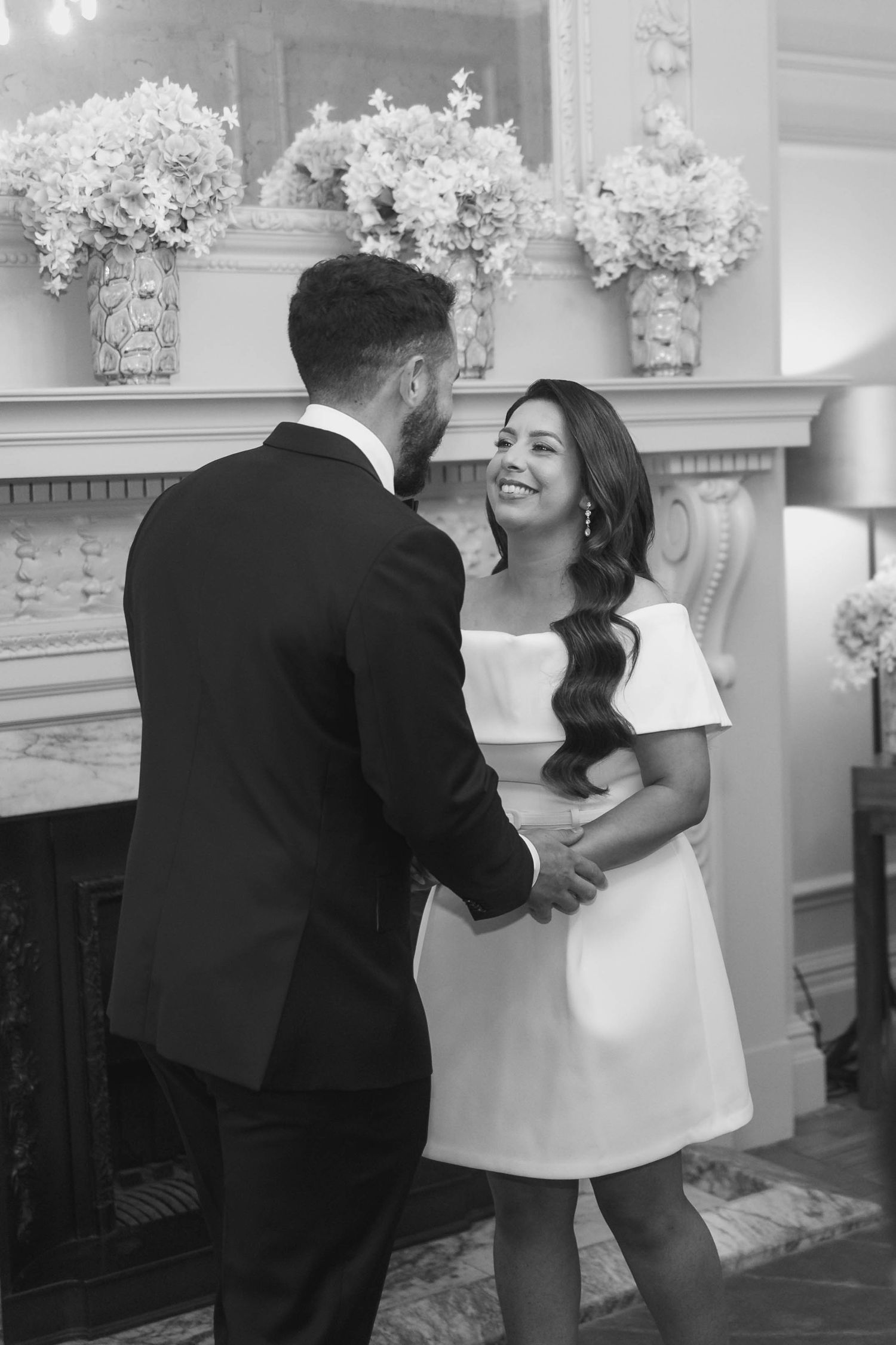 Black and white photo of a smiling bride in white mini dress holding hands with groom in tuxedo near elegant fireplace.