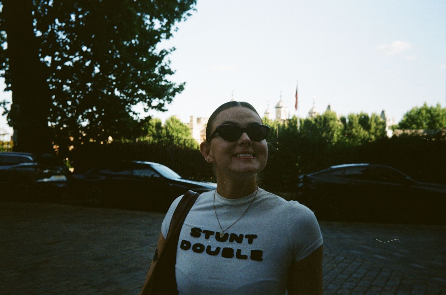 Smiling woman wearing sunglasses and a white Stunt Double t-shirt outdoors near parked cars and trees.