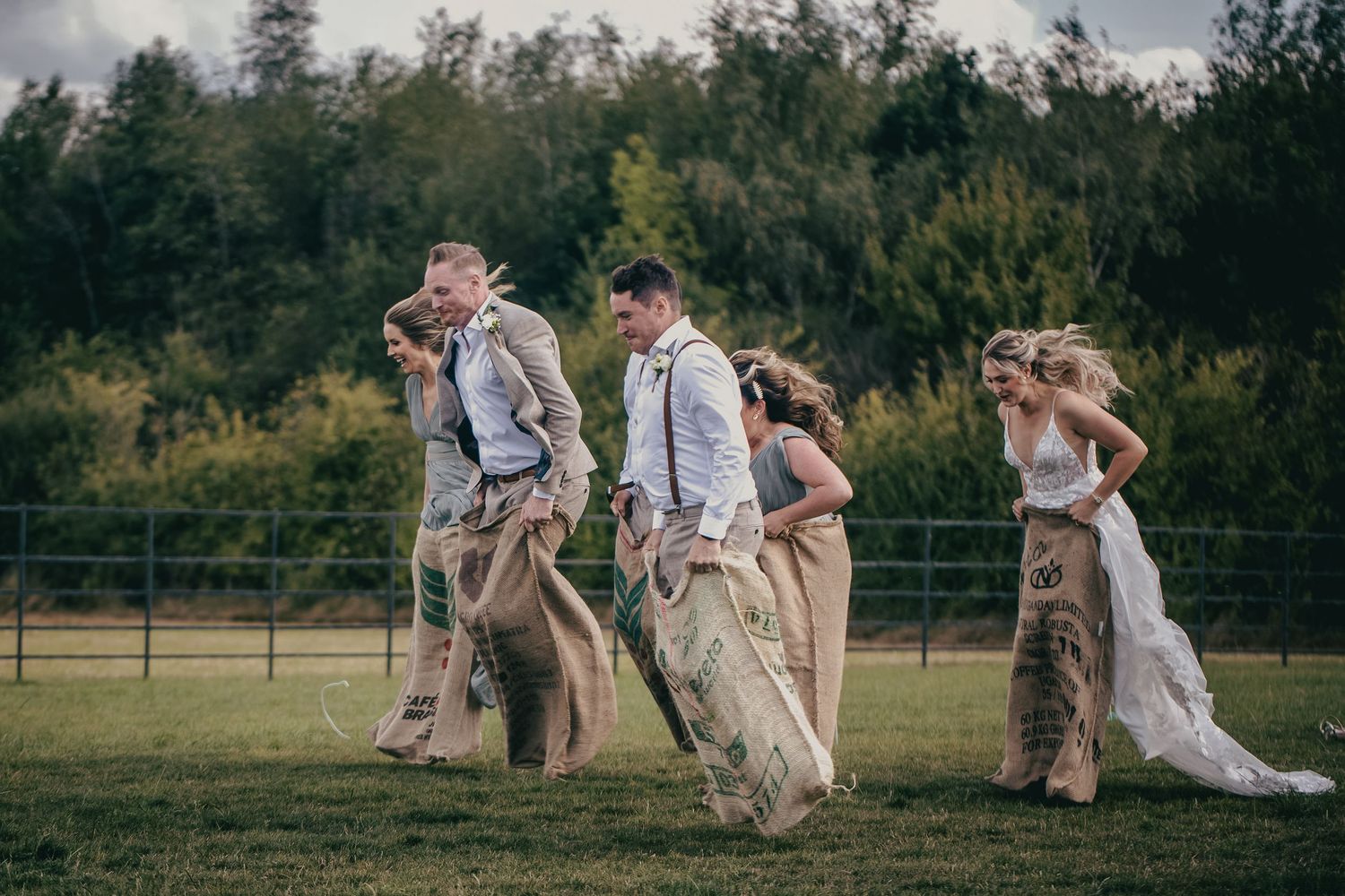 Wedding party competing in fun burlap sack race on grass field during outdoor wedding celebration.