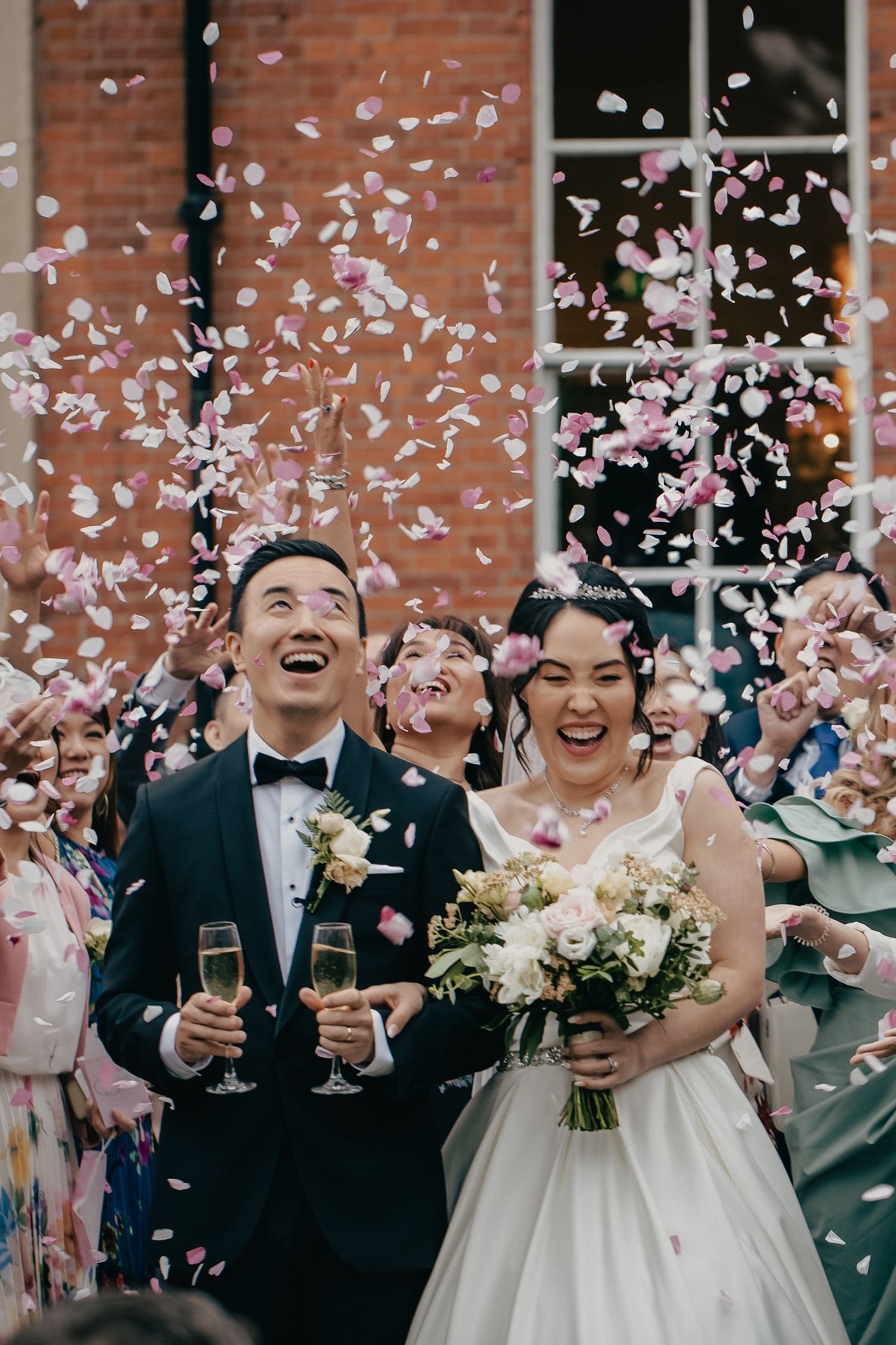Joyful newlyweds laugh as colorful confetti fills the air while holding champagne glasses after their ceremony.