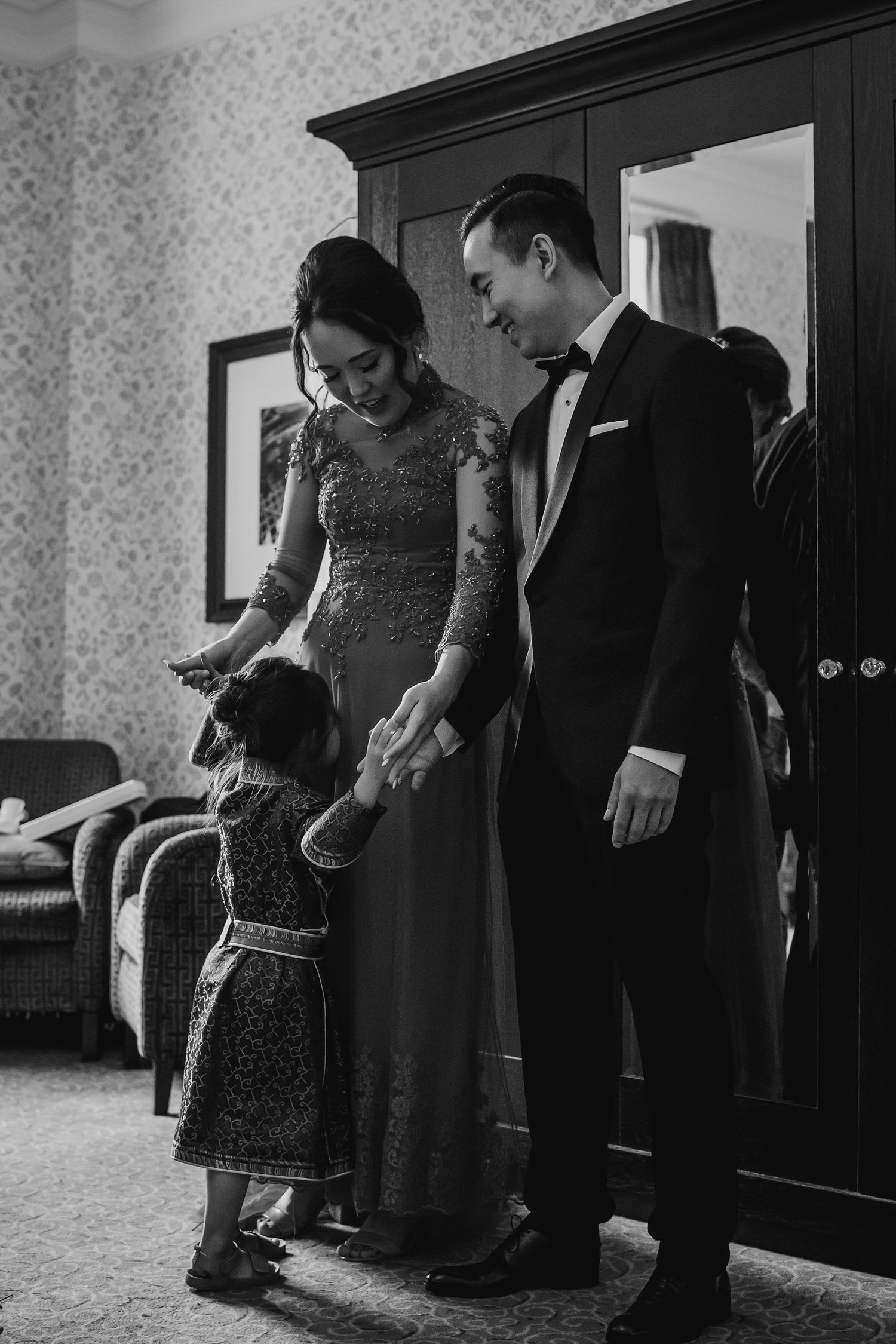 Black and white photo of a bride and groom holding hands with a small child in a hotel room.