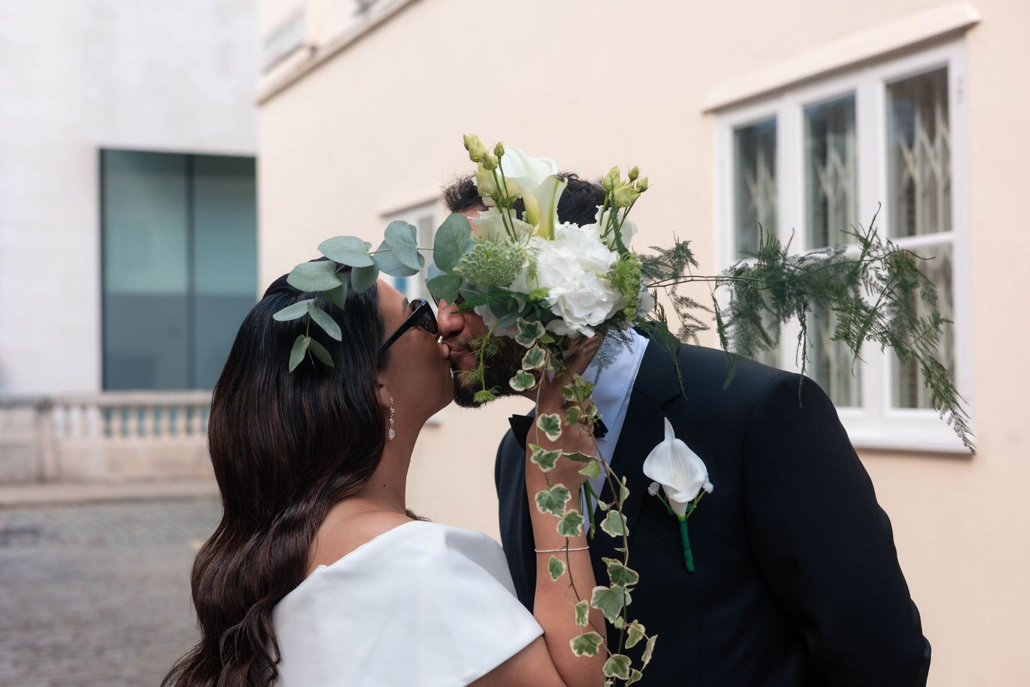 Bride and groom sharing a kiss behind a lush white floral bouquet with eucalyptus on their wedding day.