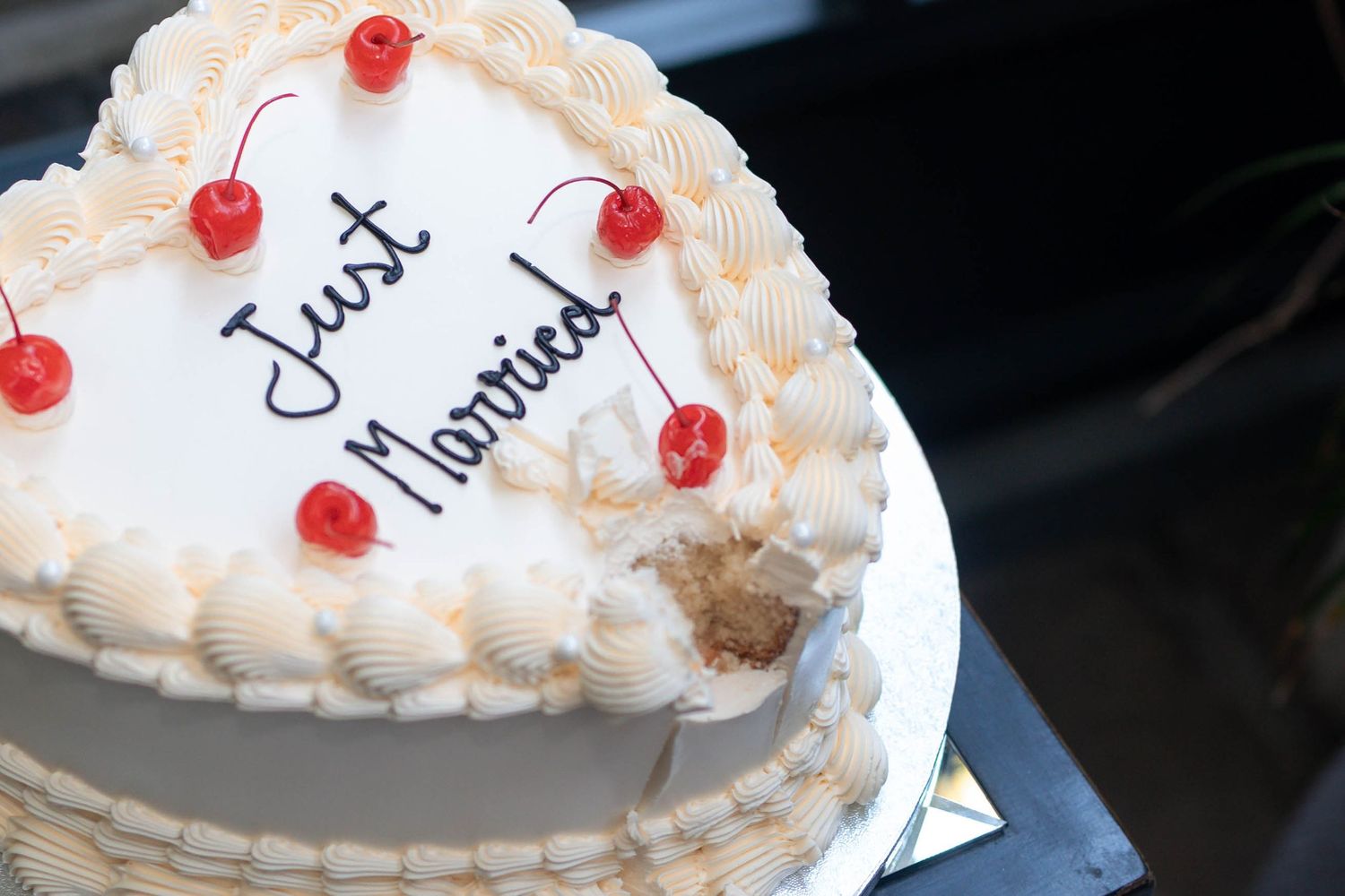Heart-shaped white wedding cake decorated with red cherries and cursive Just Married inscription on top.