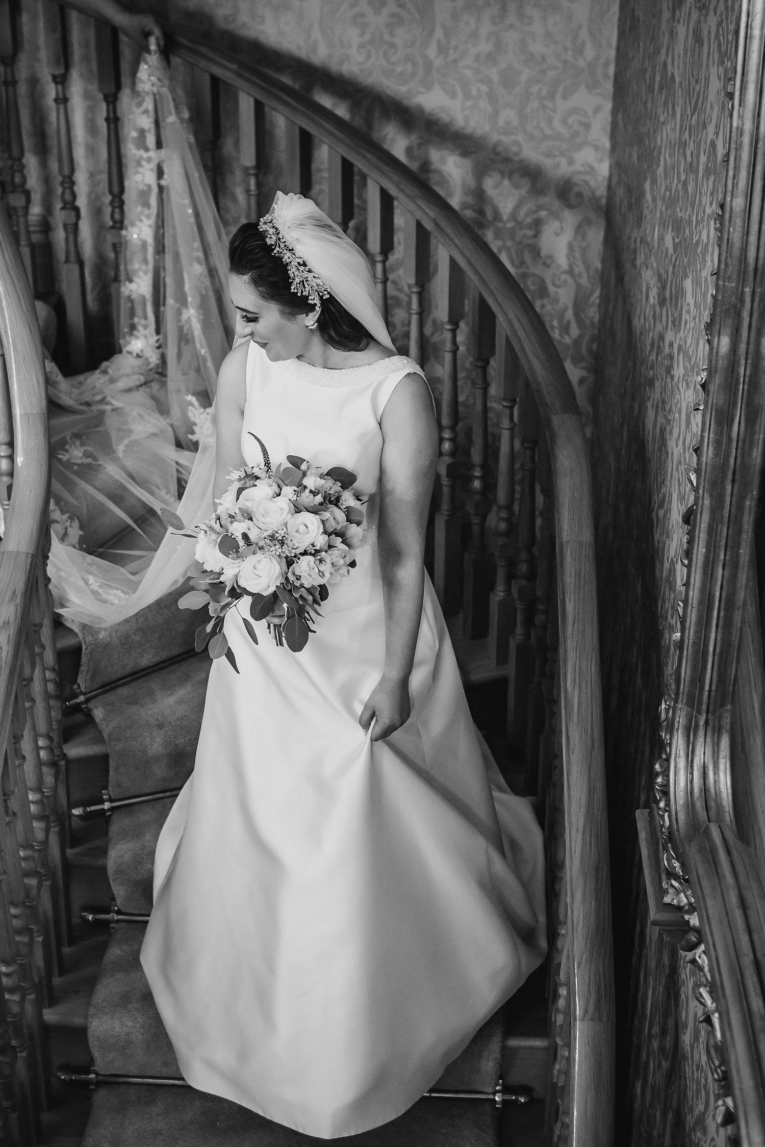 Black and white portrait of bride in classic gown holding bouquet on spiral staircase.