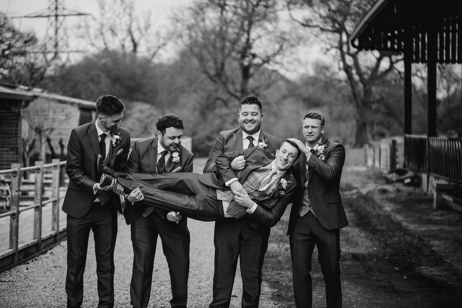 Black and white photo of groomsmen joyfully carrying groom horizontally at rustic outdoor venue.
