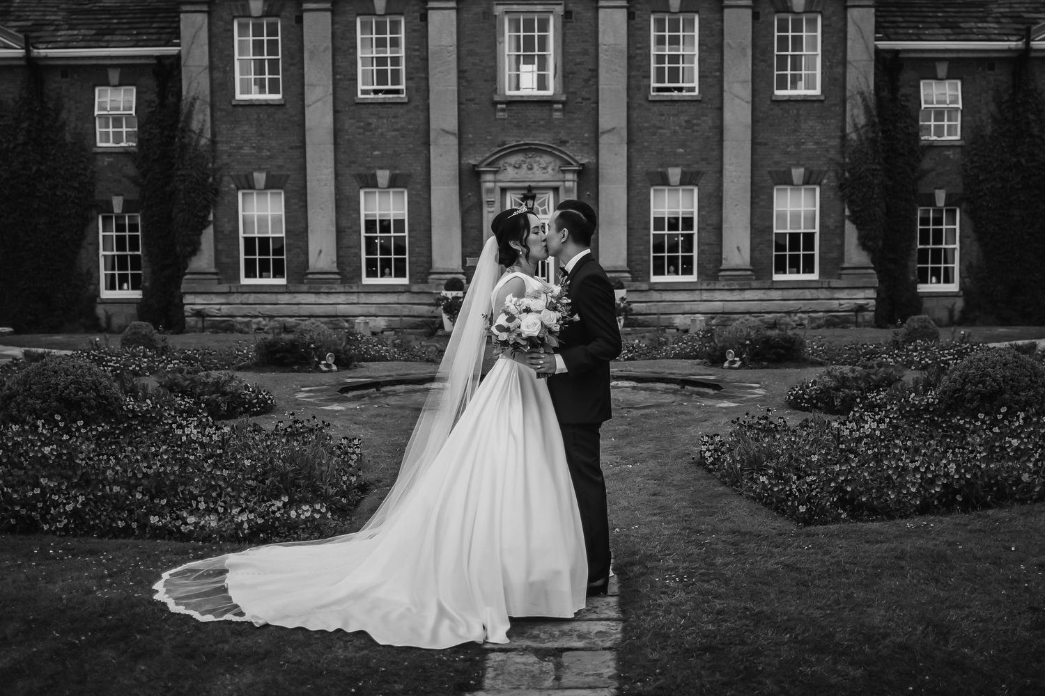 Bride and groom share a kiss outside a grand manor house with a long cathedral veil trailing behind.