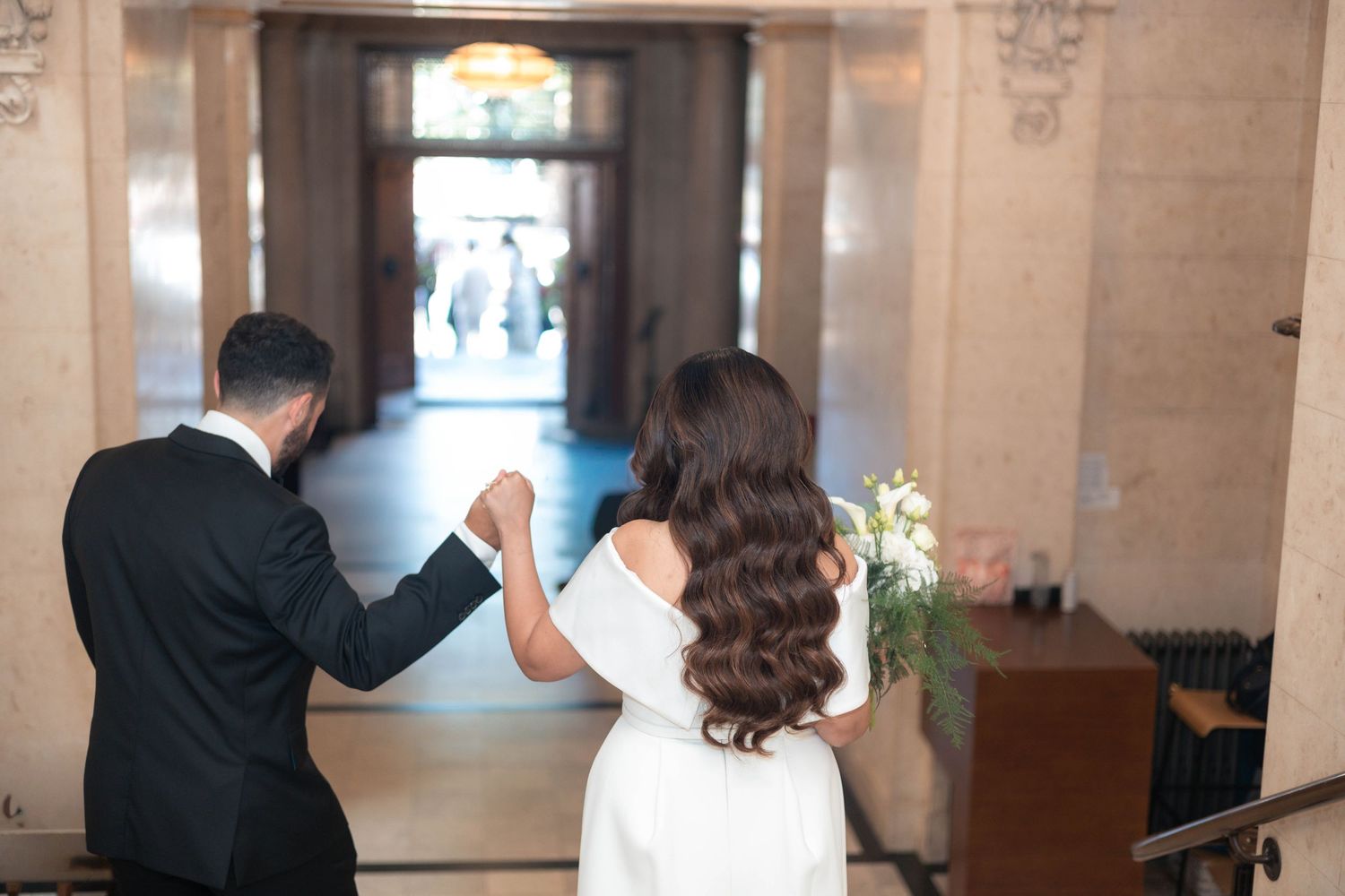 Bride and groom holding hands walking through elegant hall, woman in white dress carrying flowers.