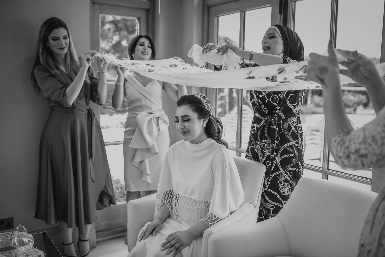 Black and white photo of bride seated as bridesmaids hold wedding veil above her.
