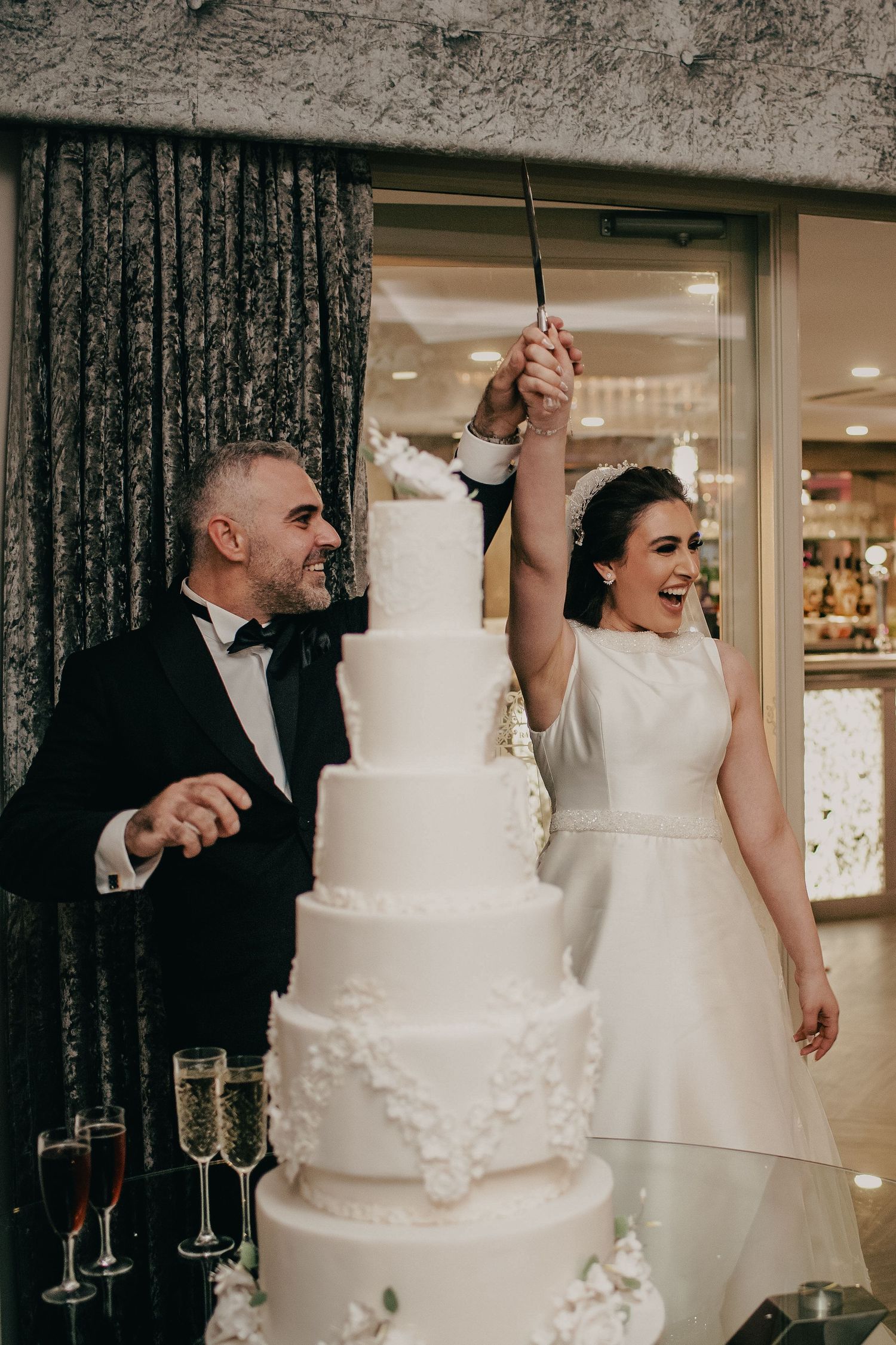 Joyful bride raising a cake cutting knife while groom smiles beside their tall white five-tier wedding cake.