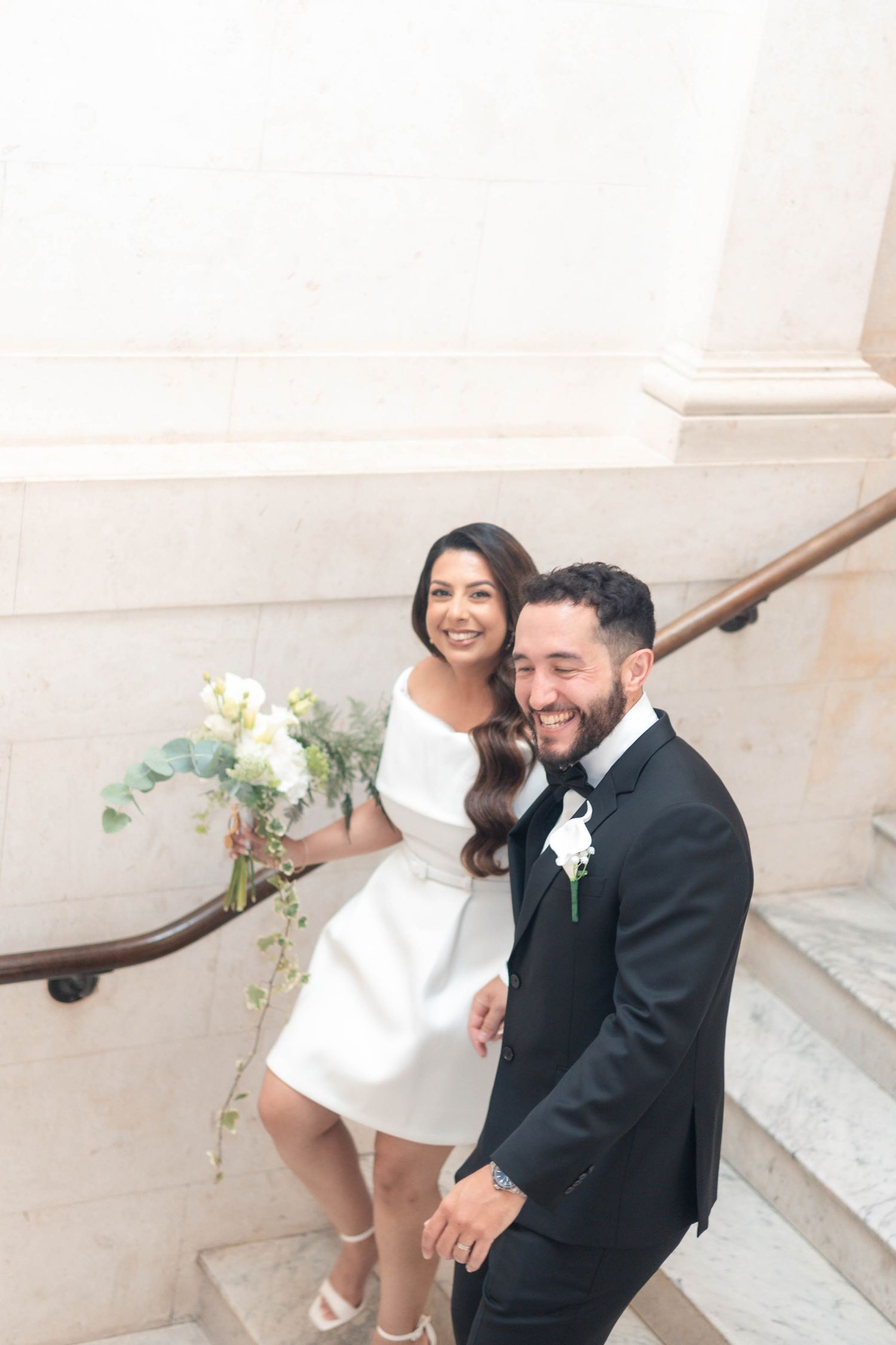 Happy couple in wedding attire smiling on marble staircase, bride in white mini dress holding bouquet, groom in black tuxedo.