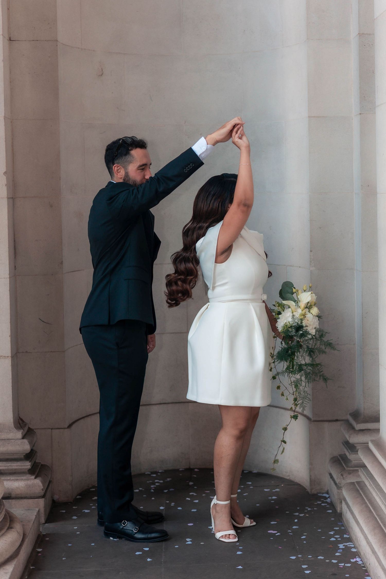 A groom in a dark suit twirls his bride in a white mini dress holding a bouquet near stone columns.