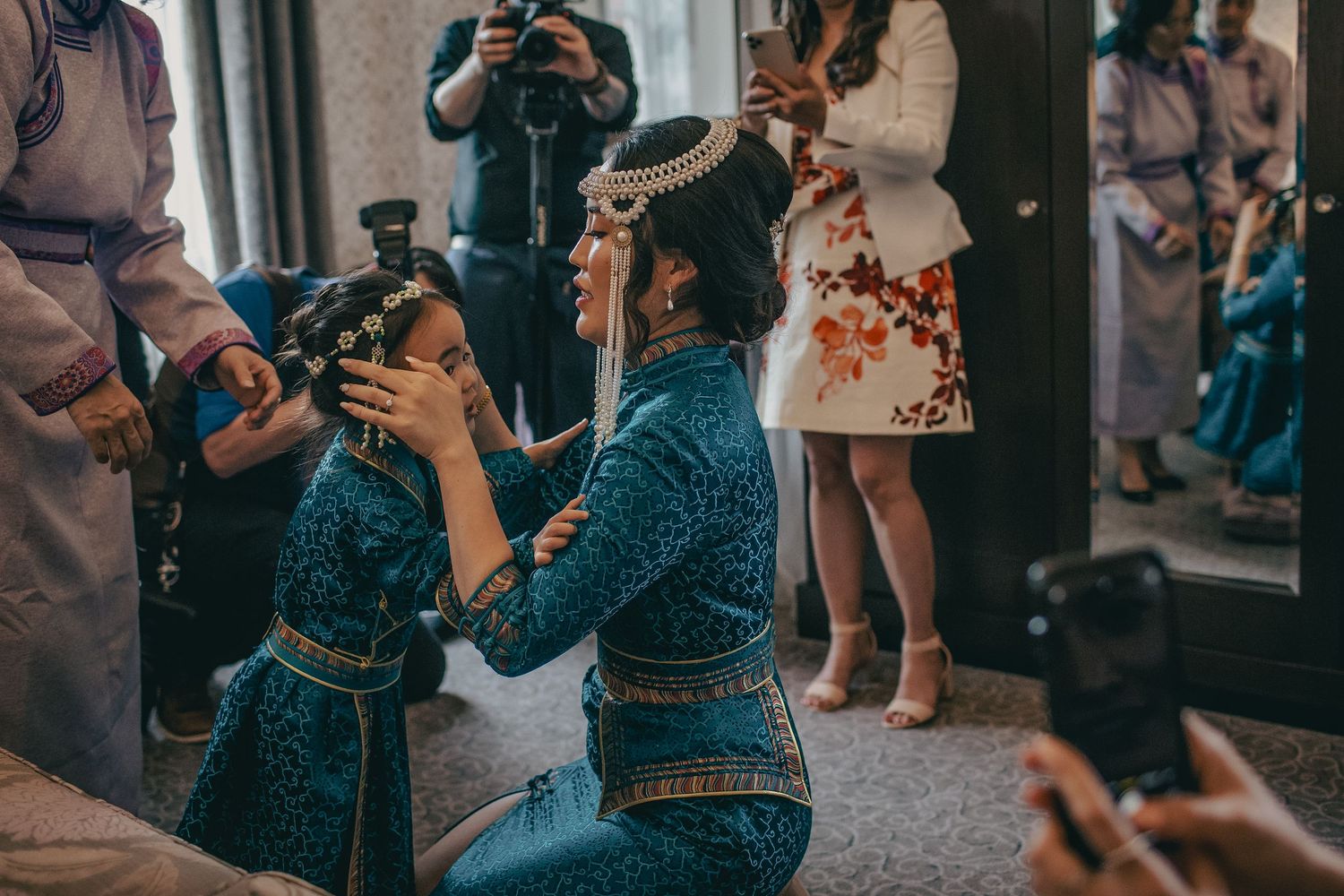 A man in traditional attire kneels to adjust a young girls floral headpiece at a wedding.