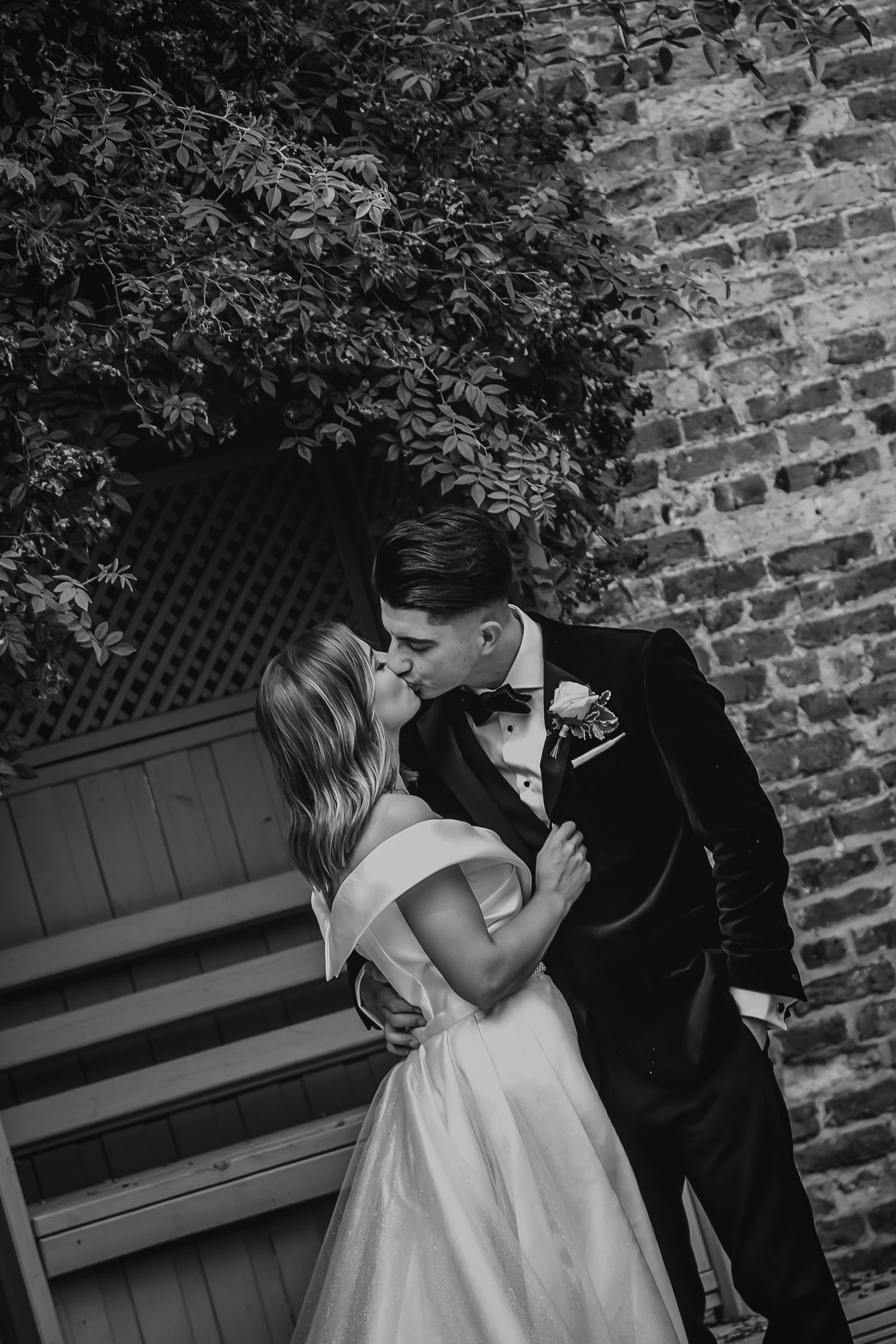 A bride and groom share a romantic kiss against a rustic brick wall in black and white.