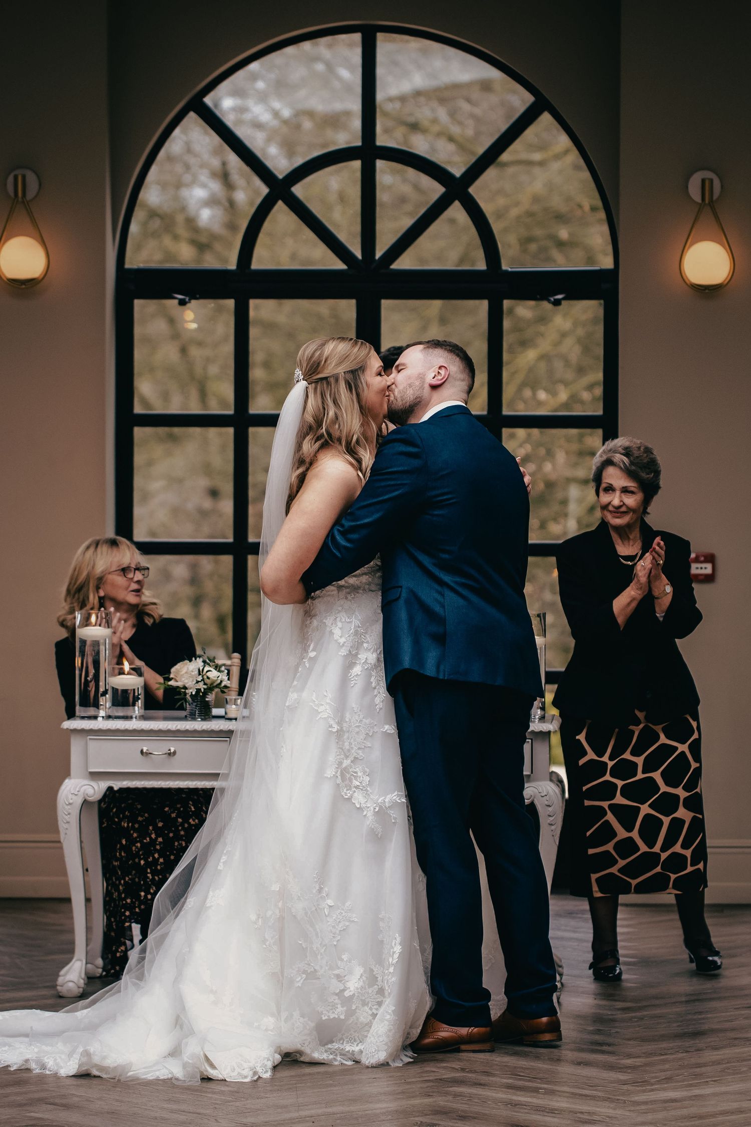 Newlyweds share first kiss before arched window as guests applaud at indoor ceremony.