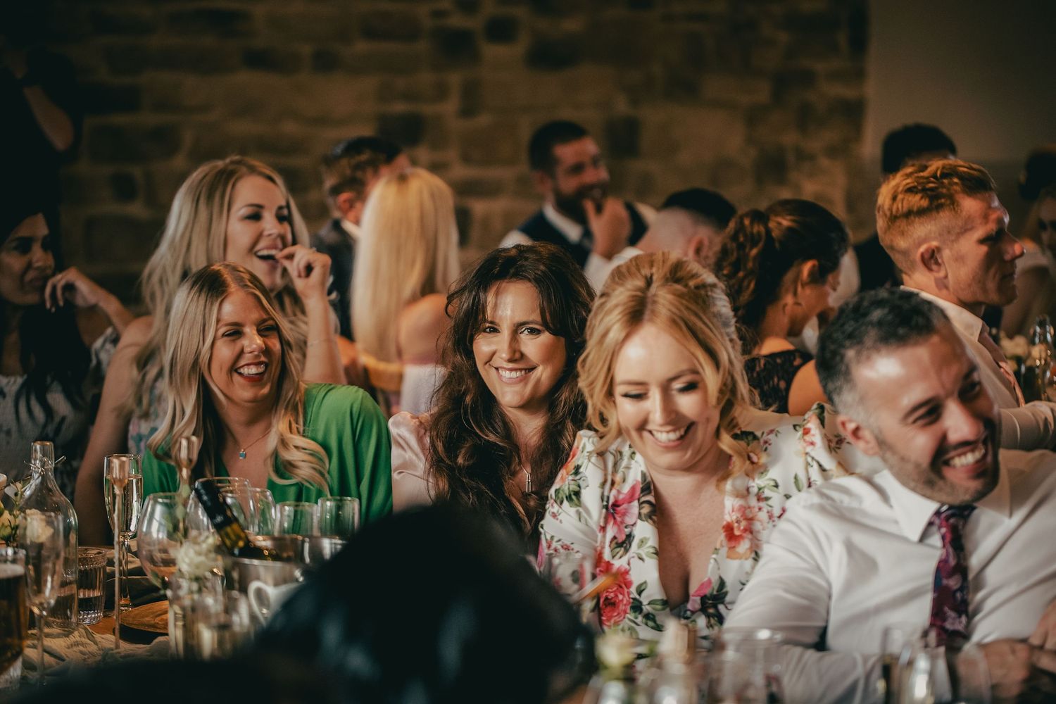 Happy wedding guests laughing together at reception dinner table in warm rustic stone barn venue.
