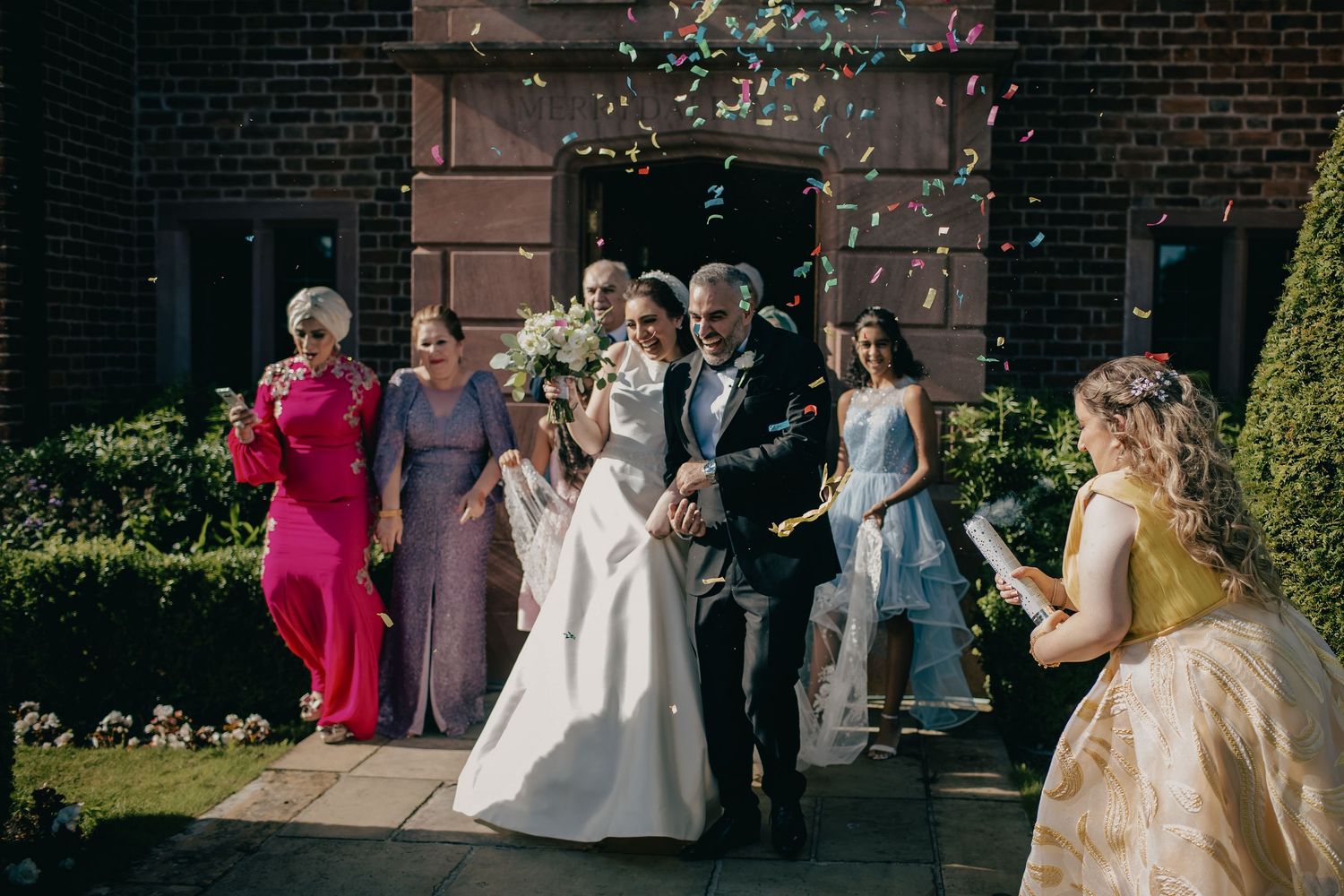 Newlyweds walk out of brick church entrance as guests toss colorful confetti in sunny outdoor setting.