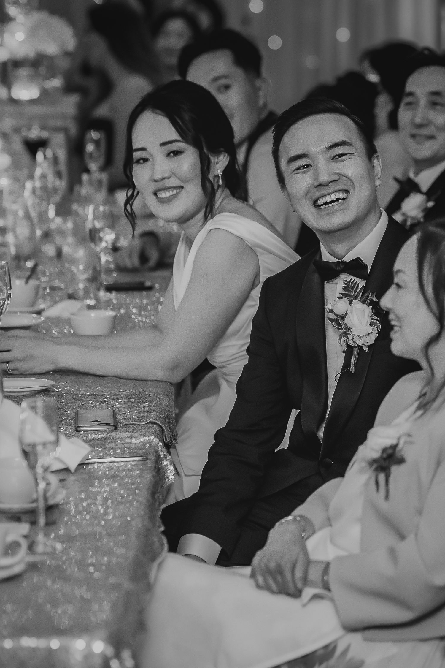 Black and white photo of smiling Asian bride and groom seated at elegant wedding reception table.