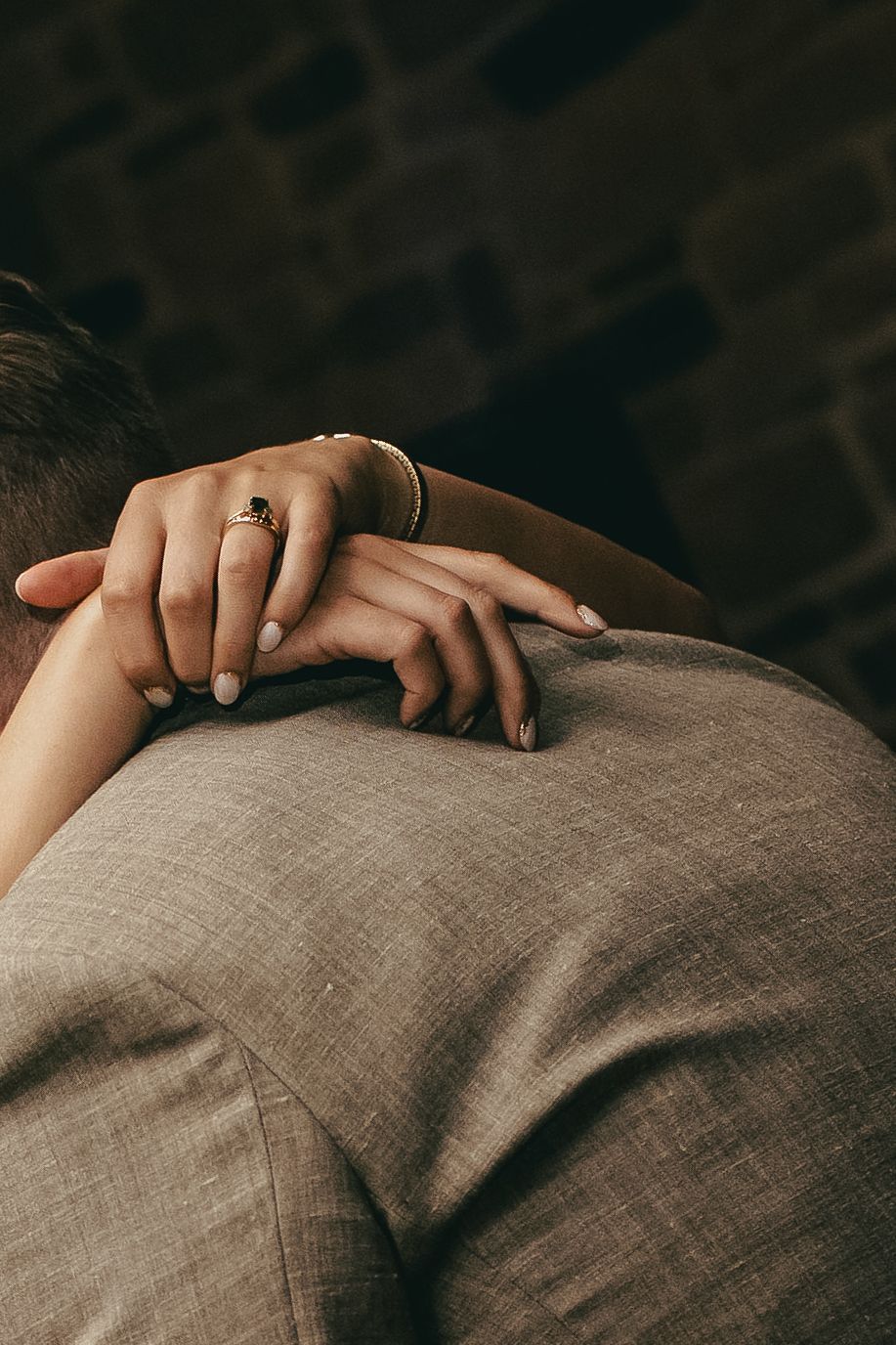 Close-up of brides hands with elegant rings resting on grooms grey suit jacket shoulder.