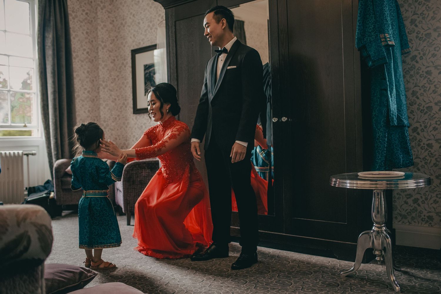 Couple in elegant red and black wedding attire stand with a young child in a hotel room.