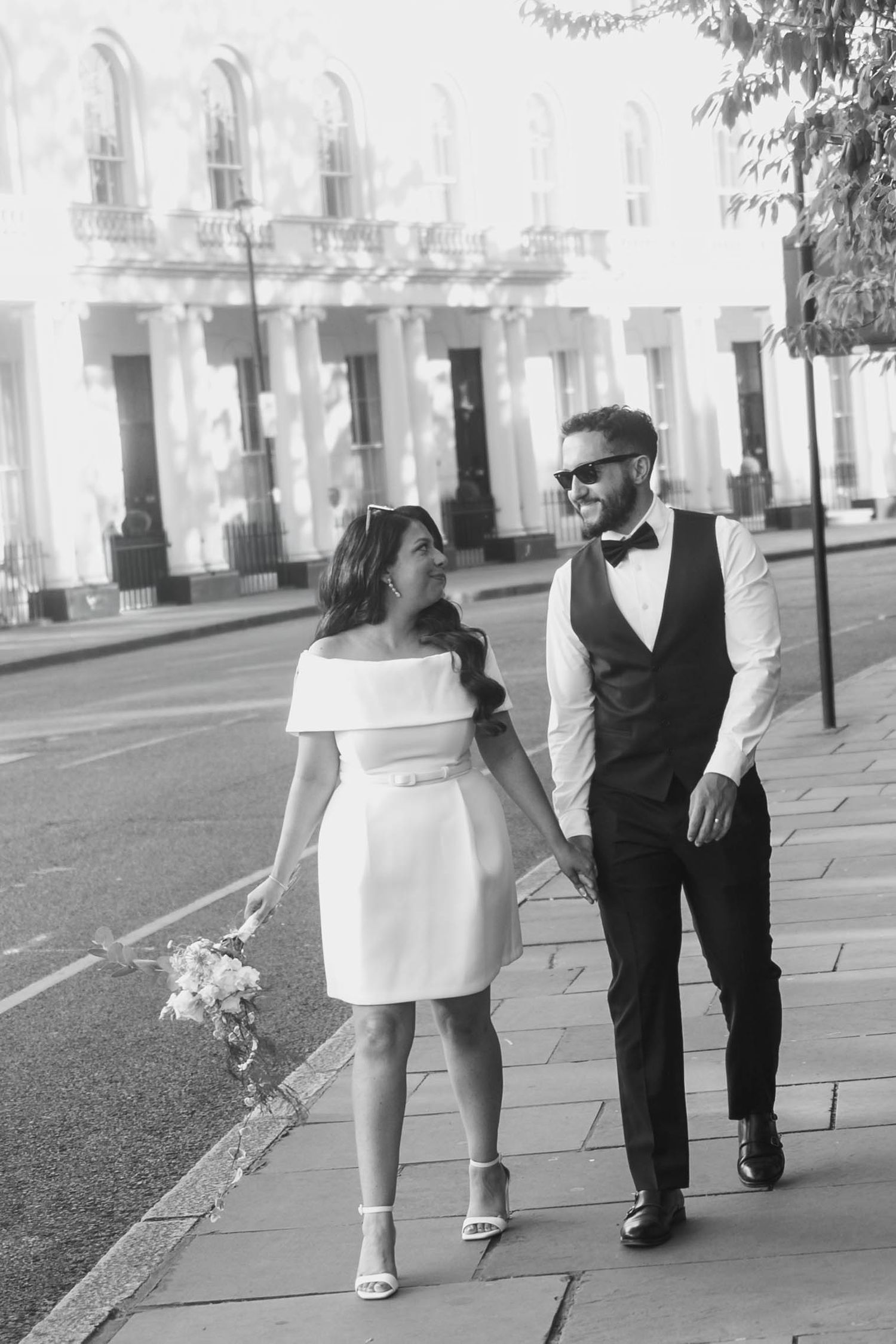 Black and white photo of a bride and groom walking hand in hand on a sunlit city sidewalk on their wedding day.