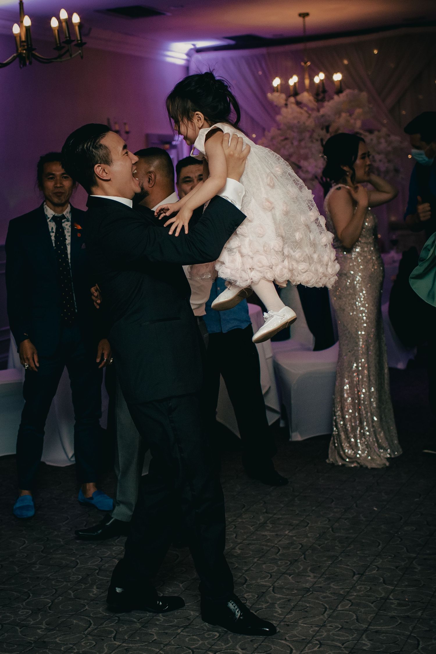 Man in black suit lifts young flower girl in white dress on colorful wedding reception dance floor.
