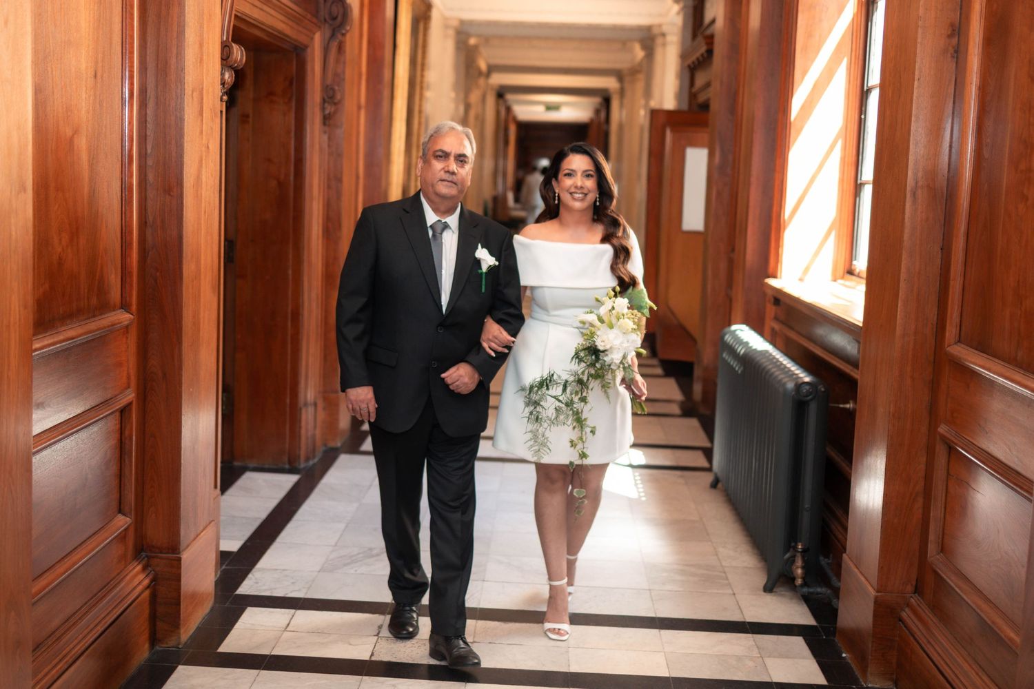 Bride in white dress walks with older man down elegant hallway with wooden doors on her wedding day.