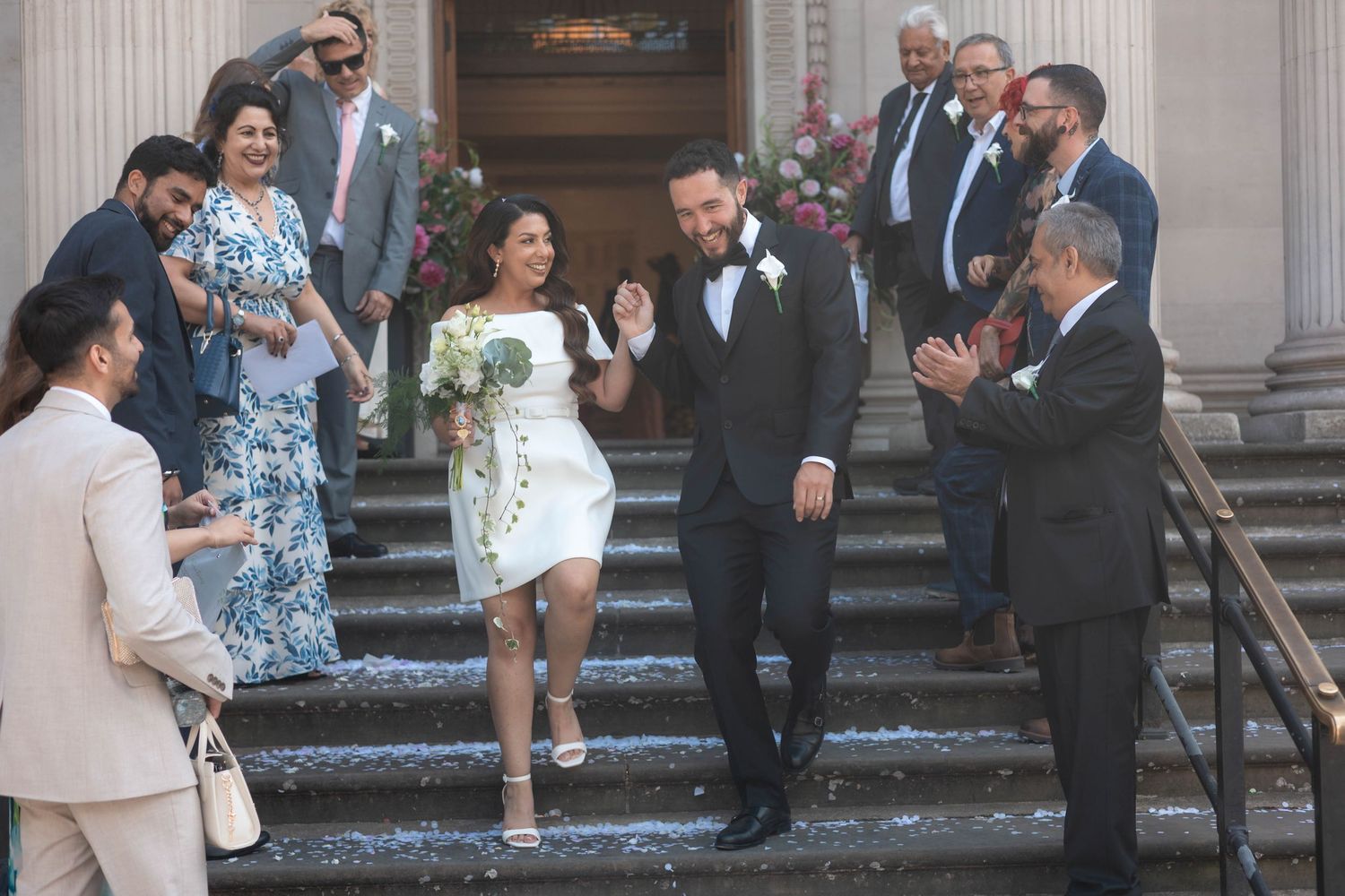 Bride and groom descend church steps in wedding attire, surrounded by applauding guests celebrating their marriage ceremony.