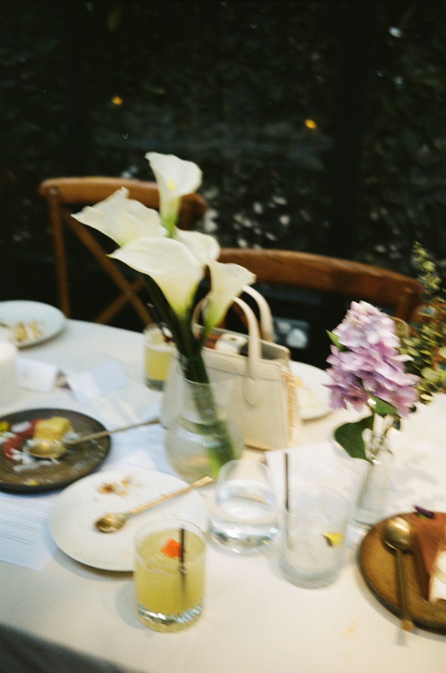 Elegant outdoor dining table with calla lilies, hydrangeas, cocktails, and white linen in a garden setting.