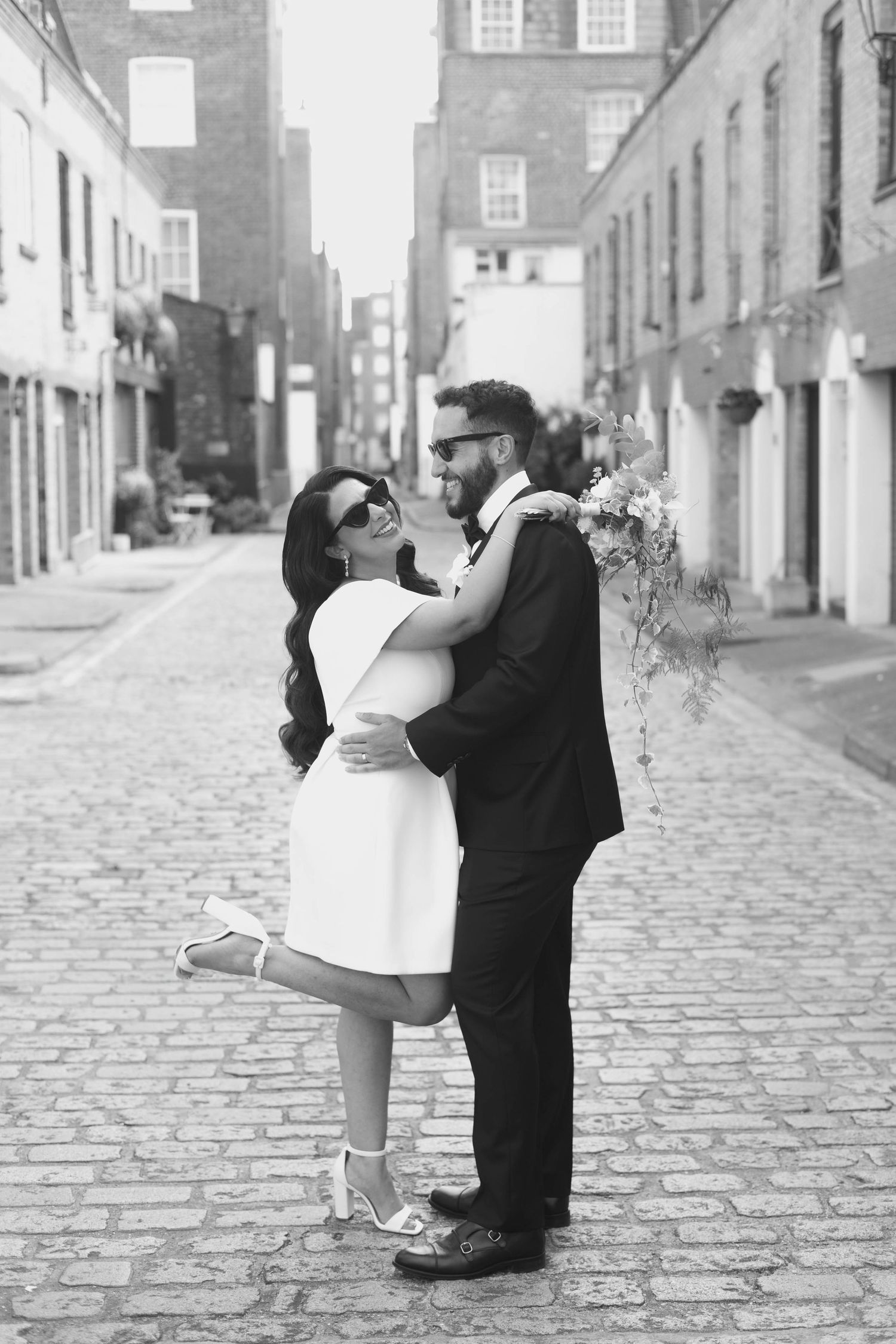 Black and white photo of a couple embracing on a cobblestone street, bride in short white dress, groom in suit holding flowers.