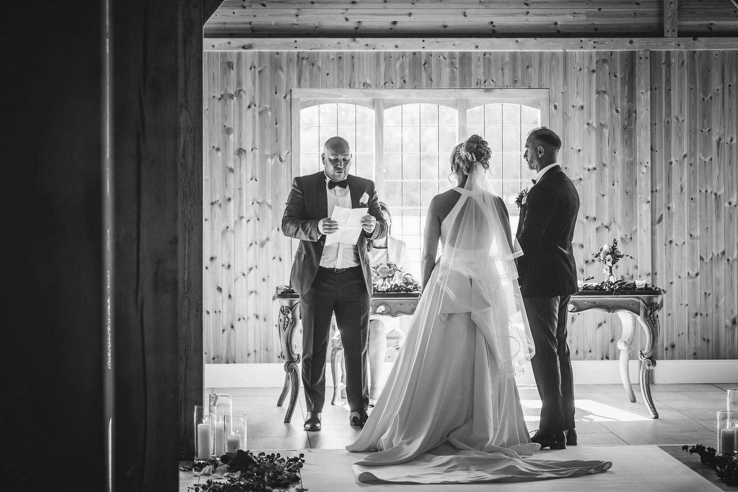 Black and white photo of bride and groom exchanging vows with officiant inside a rustic wooden barn venue.