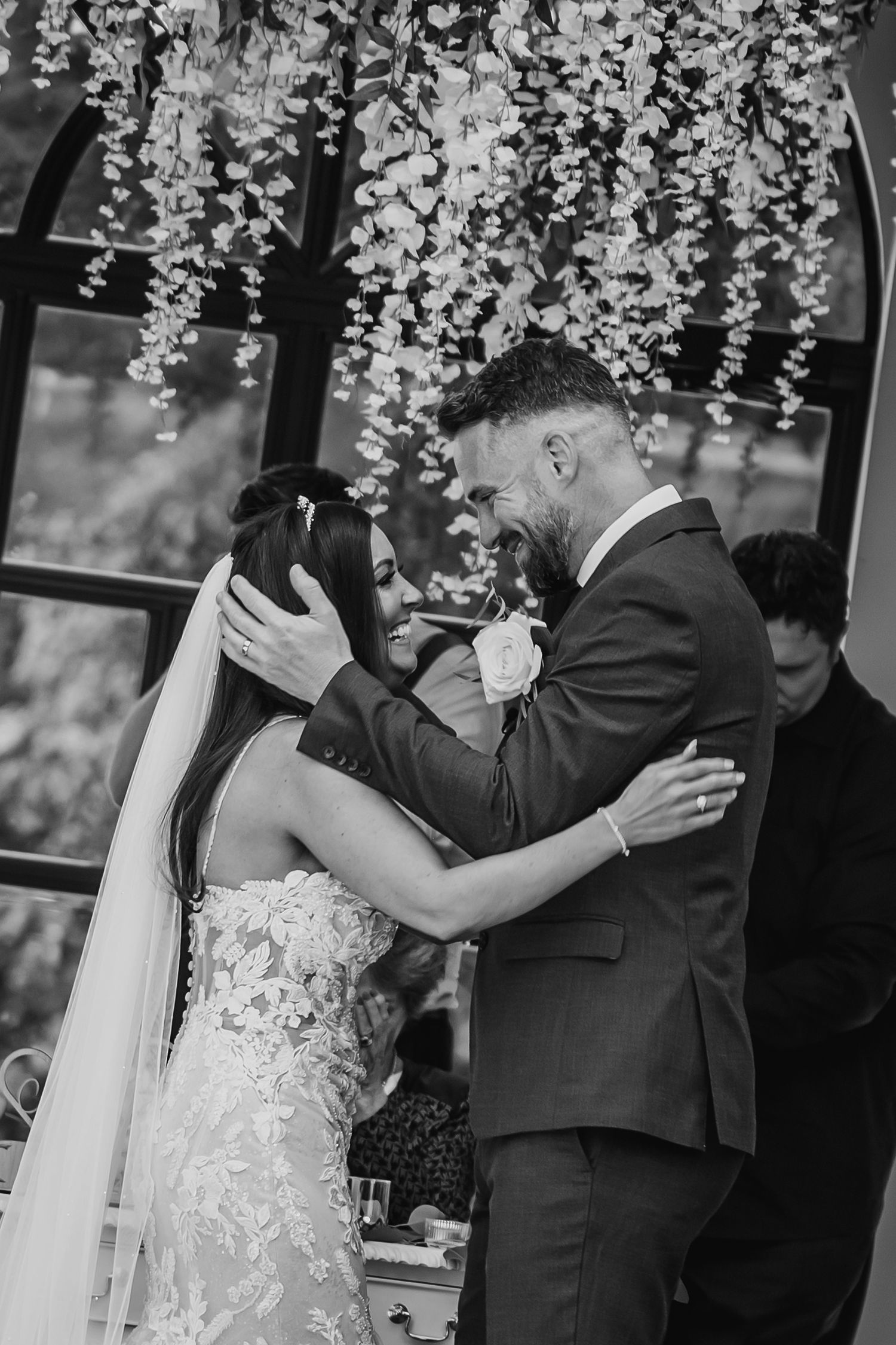 Emotional black and white close-up of bride and groom sharing first kiss under hanging floral decorations.
