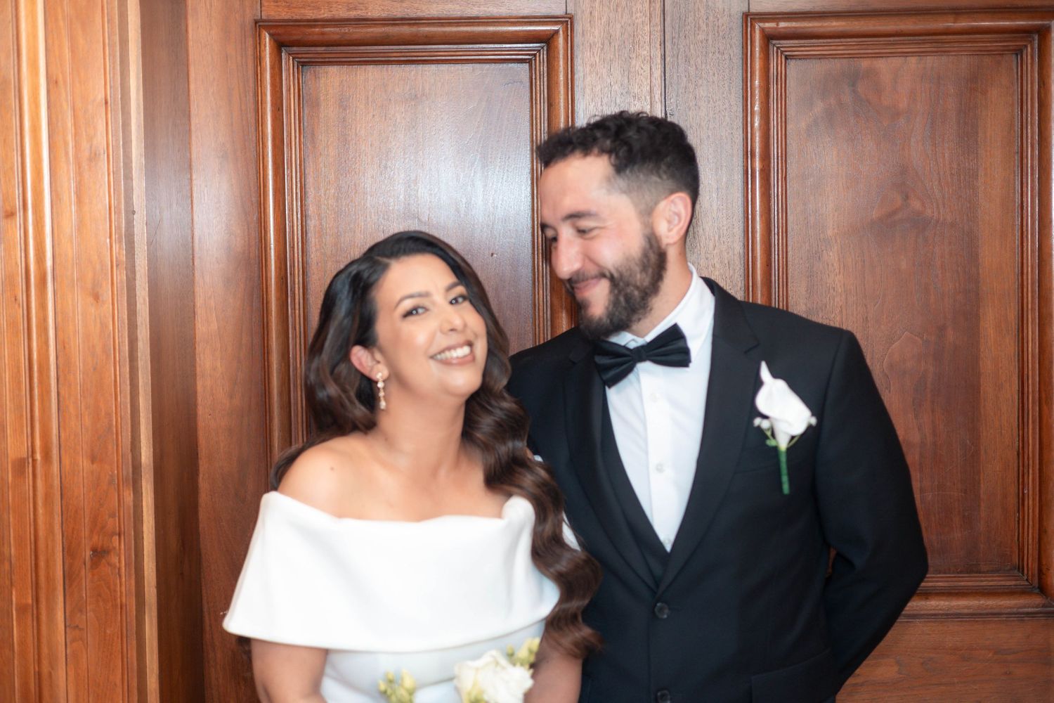 Happy couple in wedding attire smiling at each other, bride in white dress, groom in black tuxedo with boutonniere.