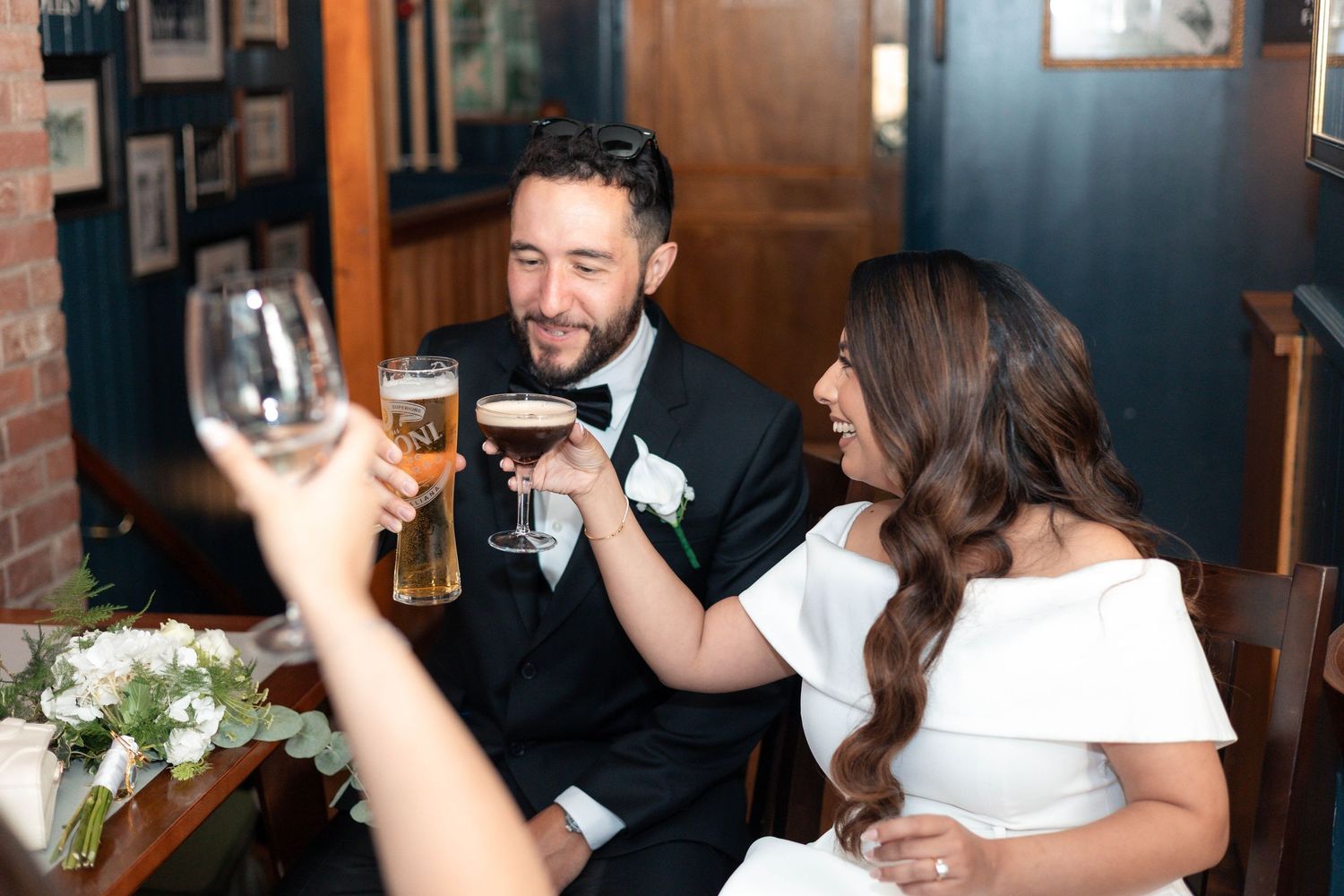 Bride and groom toast with beer and wine at wedding reception, smiling joyfully at elegant venue.