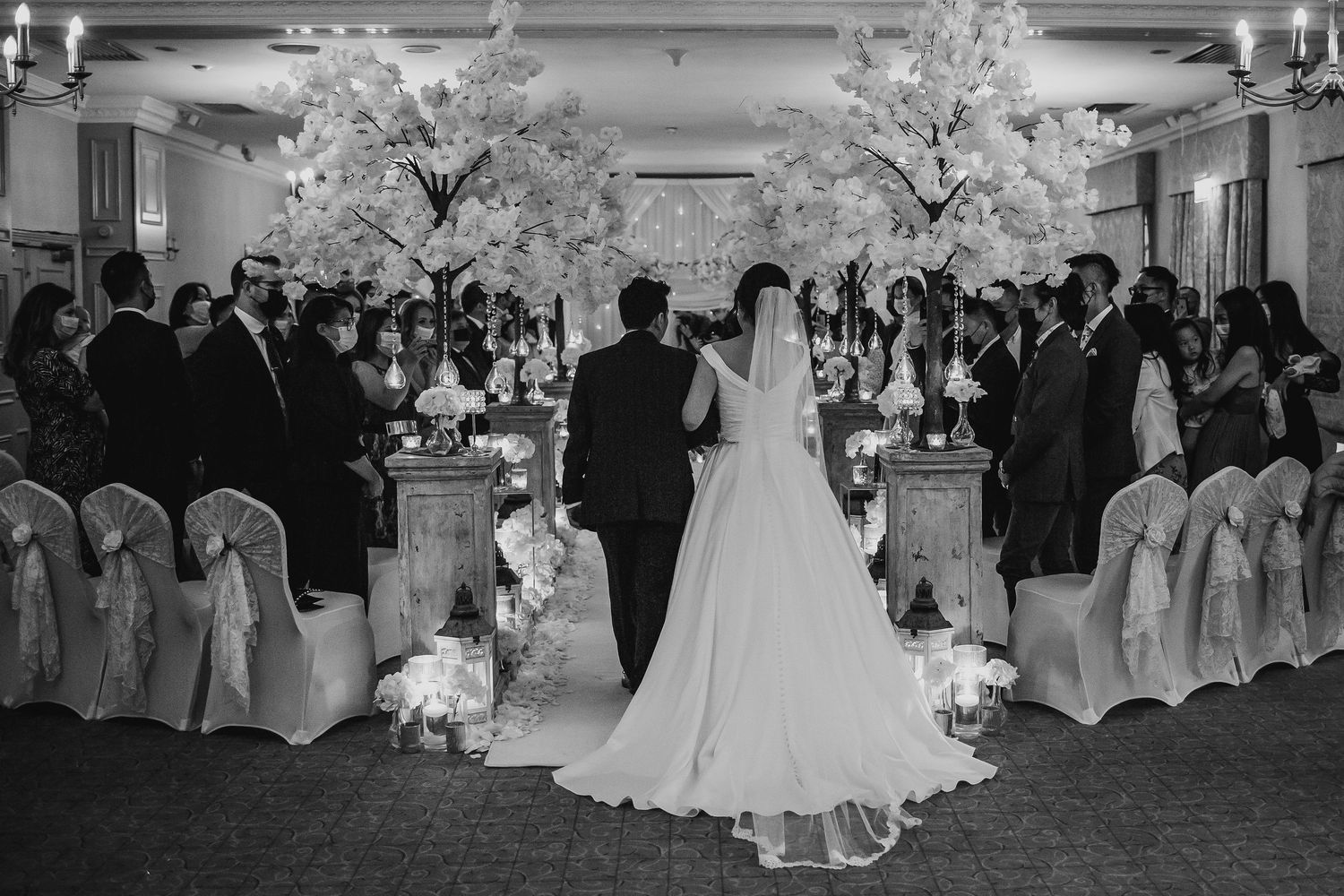 Bride walks down the aisle flanked by white blossom trees and candles in an elegant wedding ceremony.