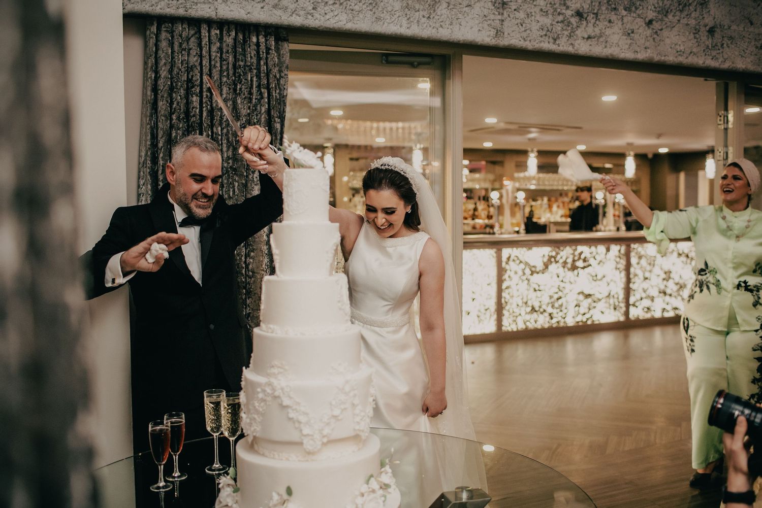 Bride and groom celebrating beside their tall white wedding cake as guests cheer in the background.