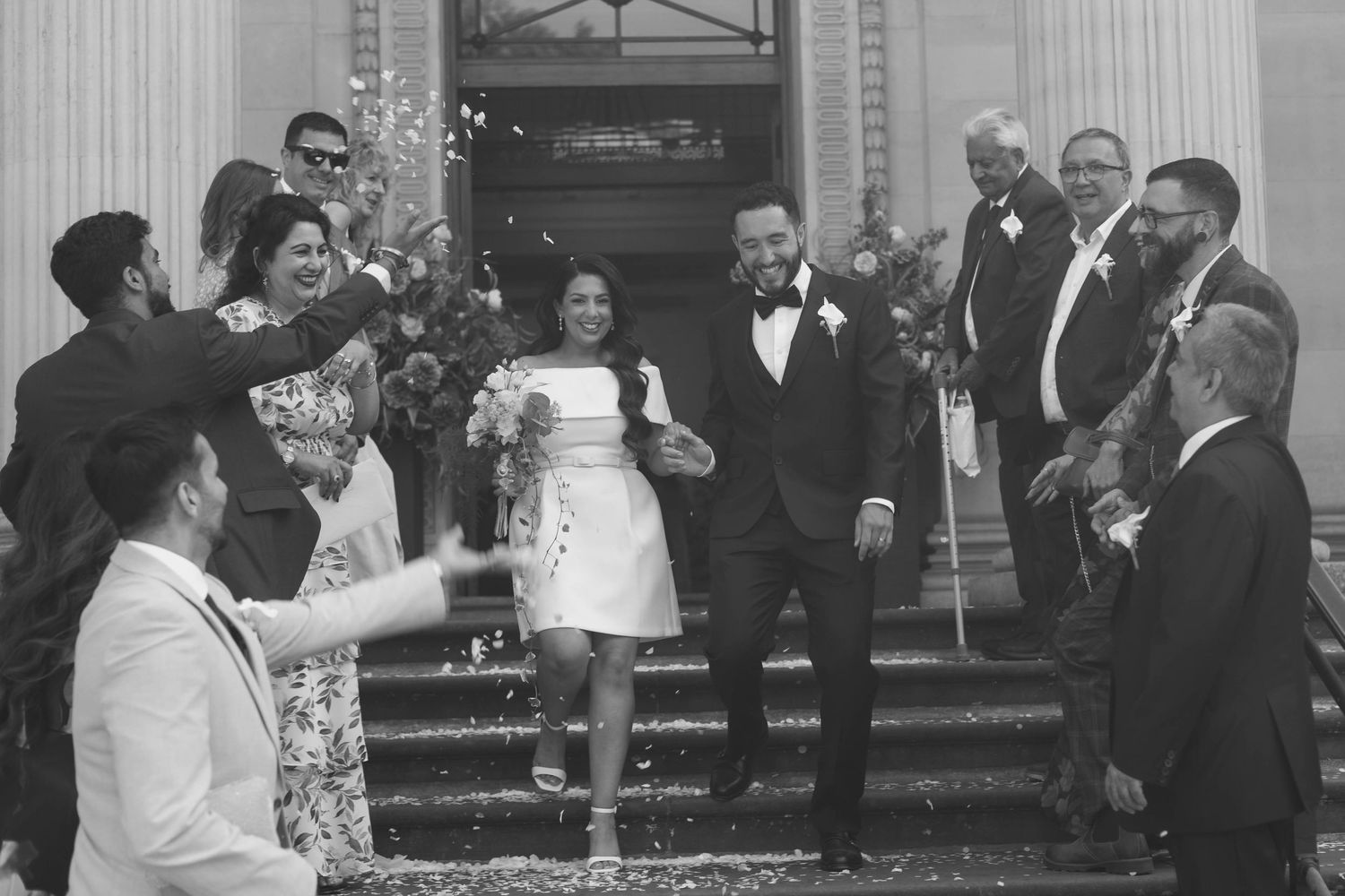 Black and white photo of a joyful newlywed couple exiting a building surrounded by cheering guests throwing confetti.